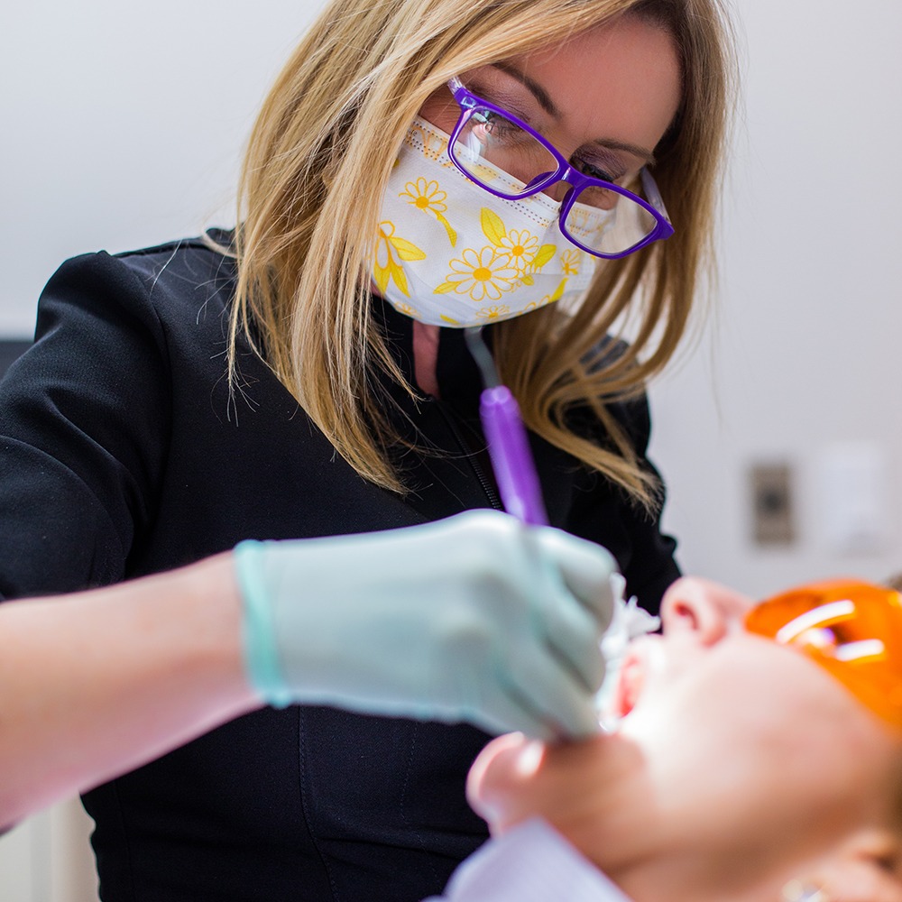 A person wearing a mask and glasses performs a dental procedure on another person in a medical setting.