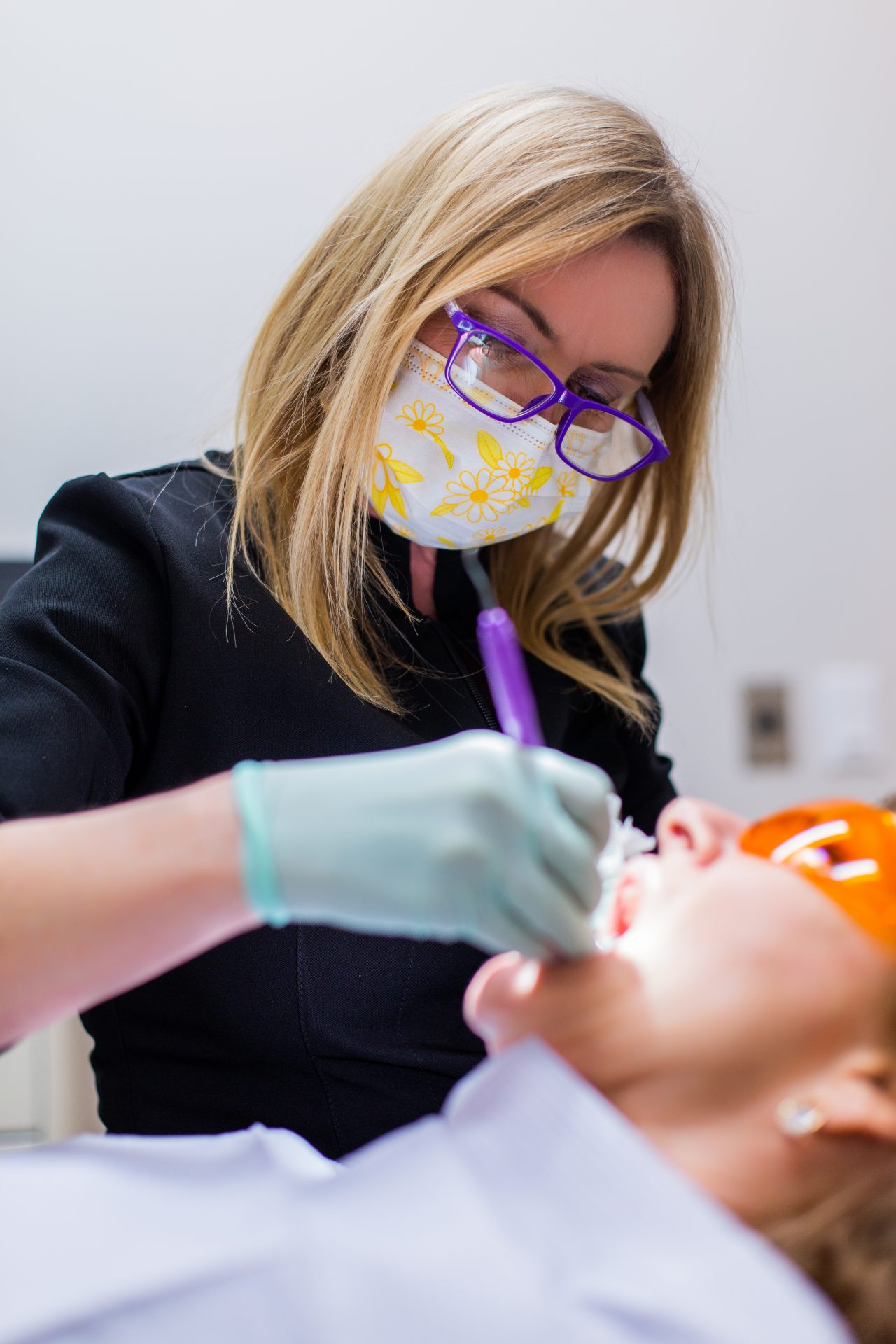 A person wearing a mask and glasses performs a dental procedure on another person, who is reclining with protective eyewear.