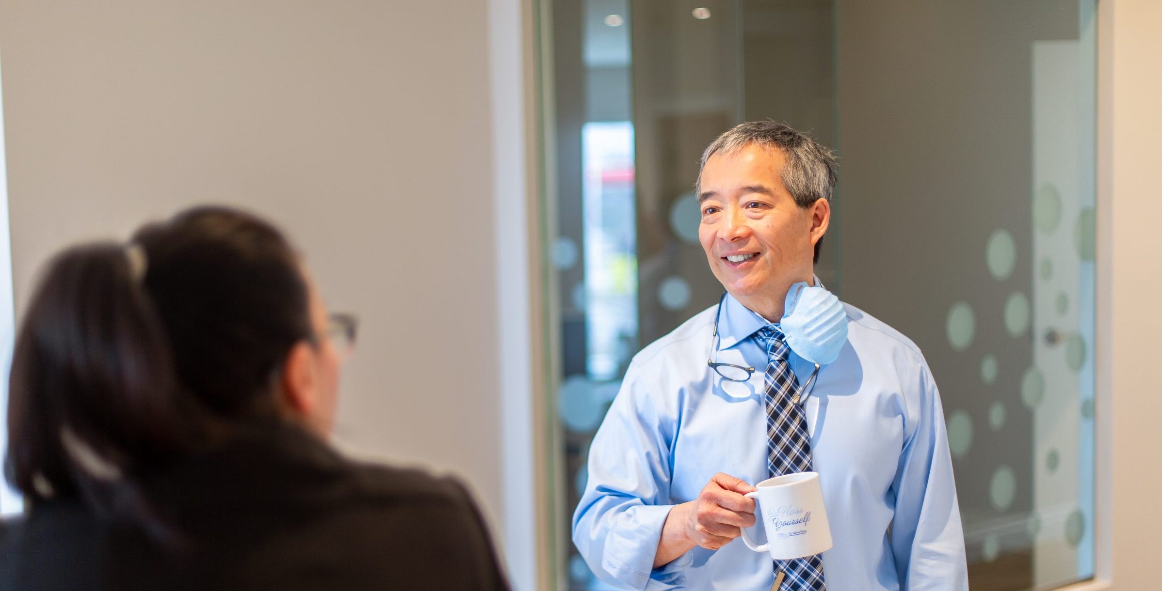 Two people in a bright office setting, smiling. One holds a coffee mug and wears a blue shirt and tie.