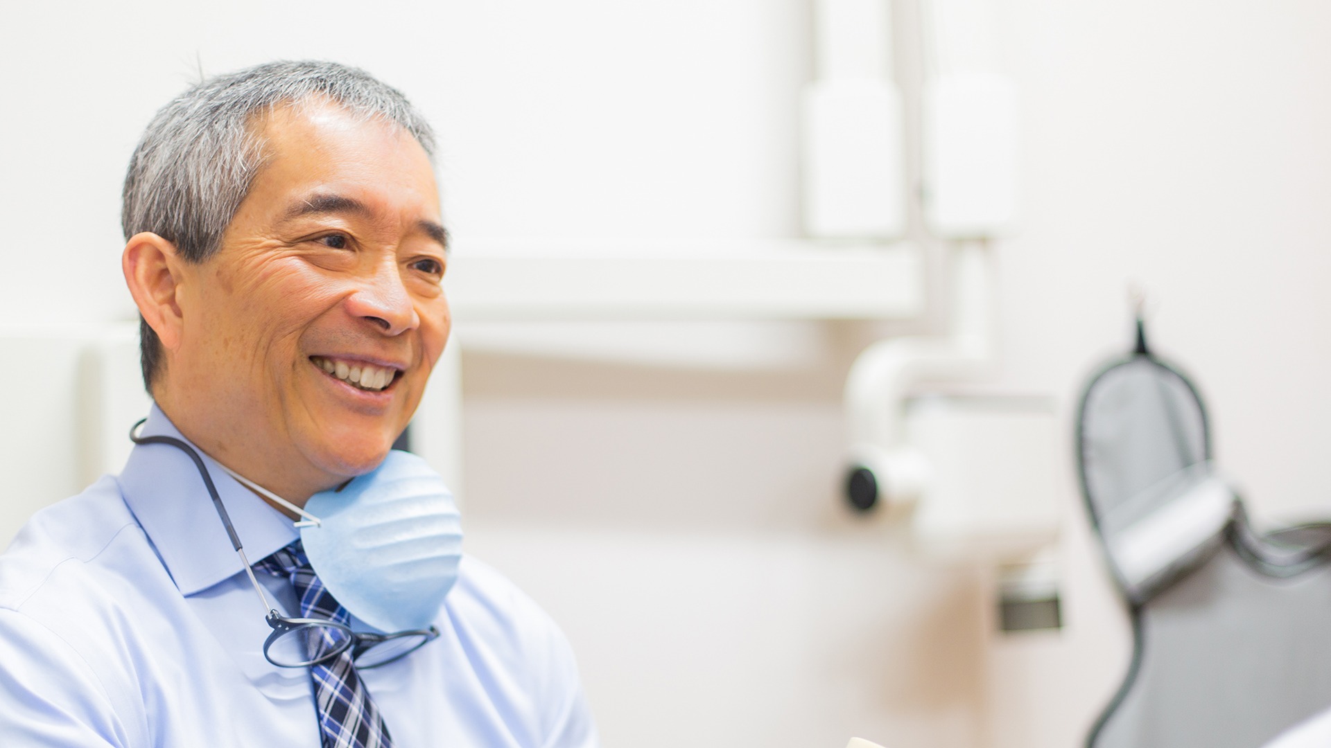 A smiling person with grey hair wears a blue shirt, glasses, and a mask around the neck in a clinical setting.