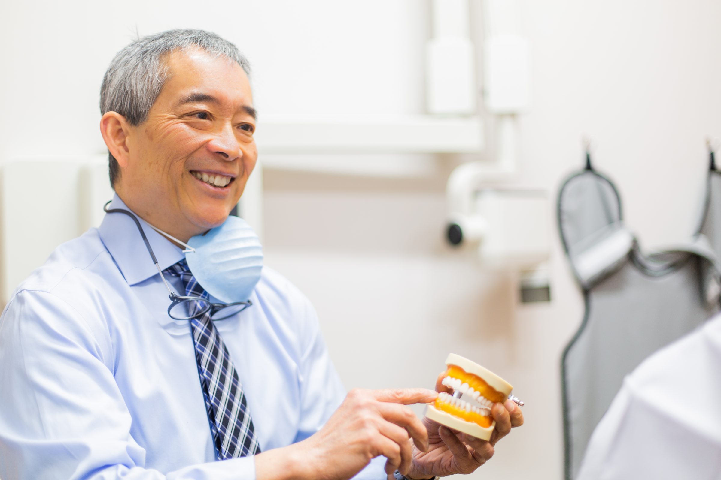 A person in a dental clinic smiles while holding a dental model, wearing a mask and glasses around their neck.