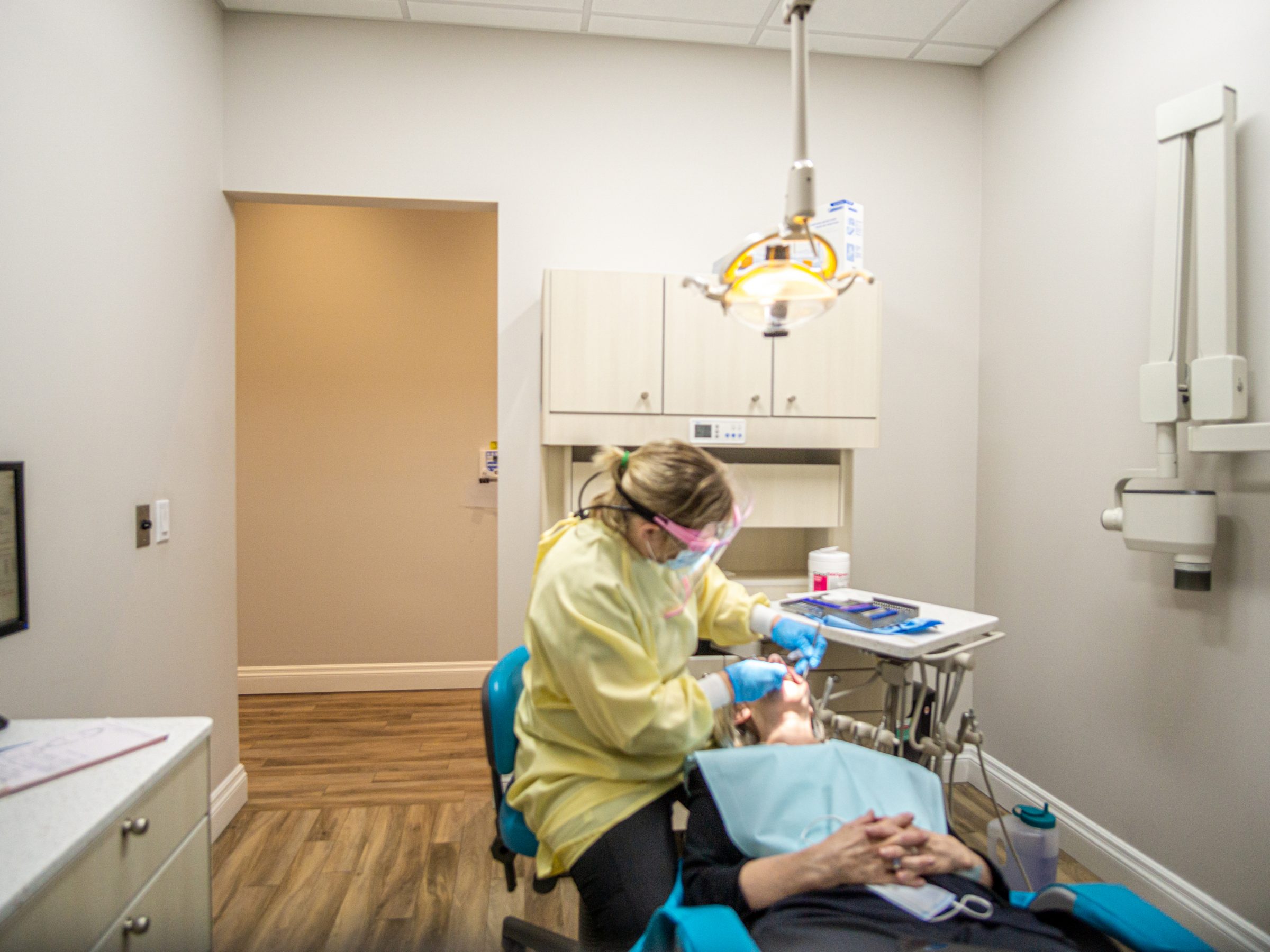 A dental office scene with a person in protective gear examining a patient, surrounded by dental equipment and a bright ceiling light.