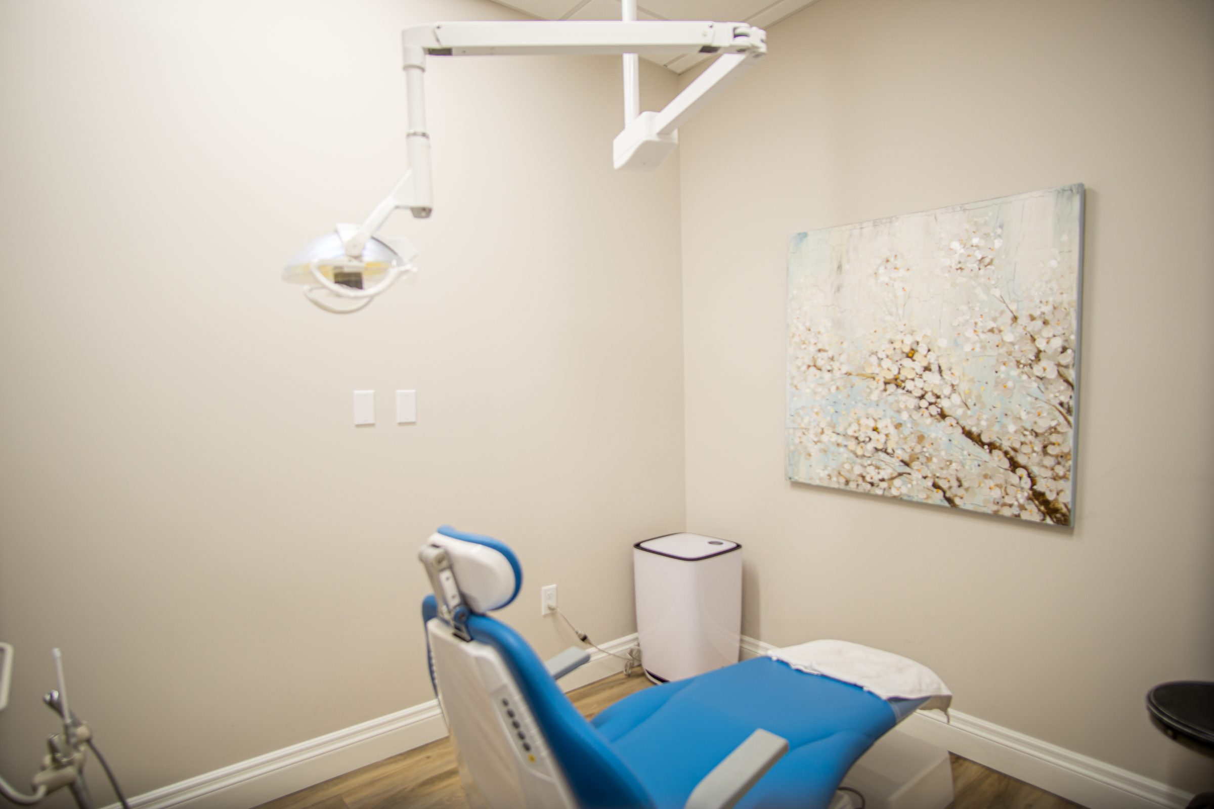 Dental office with blue chair, overhead light, abstract floral painting on wall, and wooden floor. White walls and a waste bin are visible.