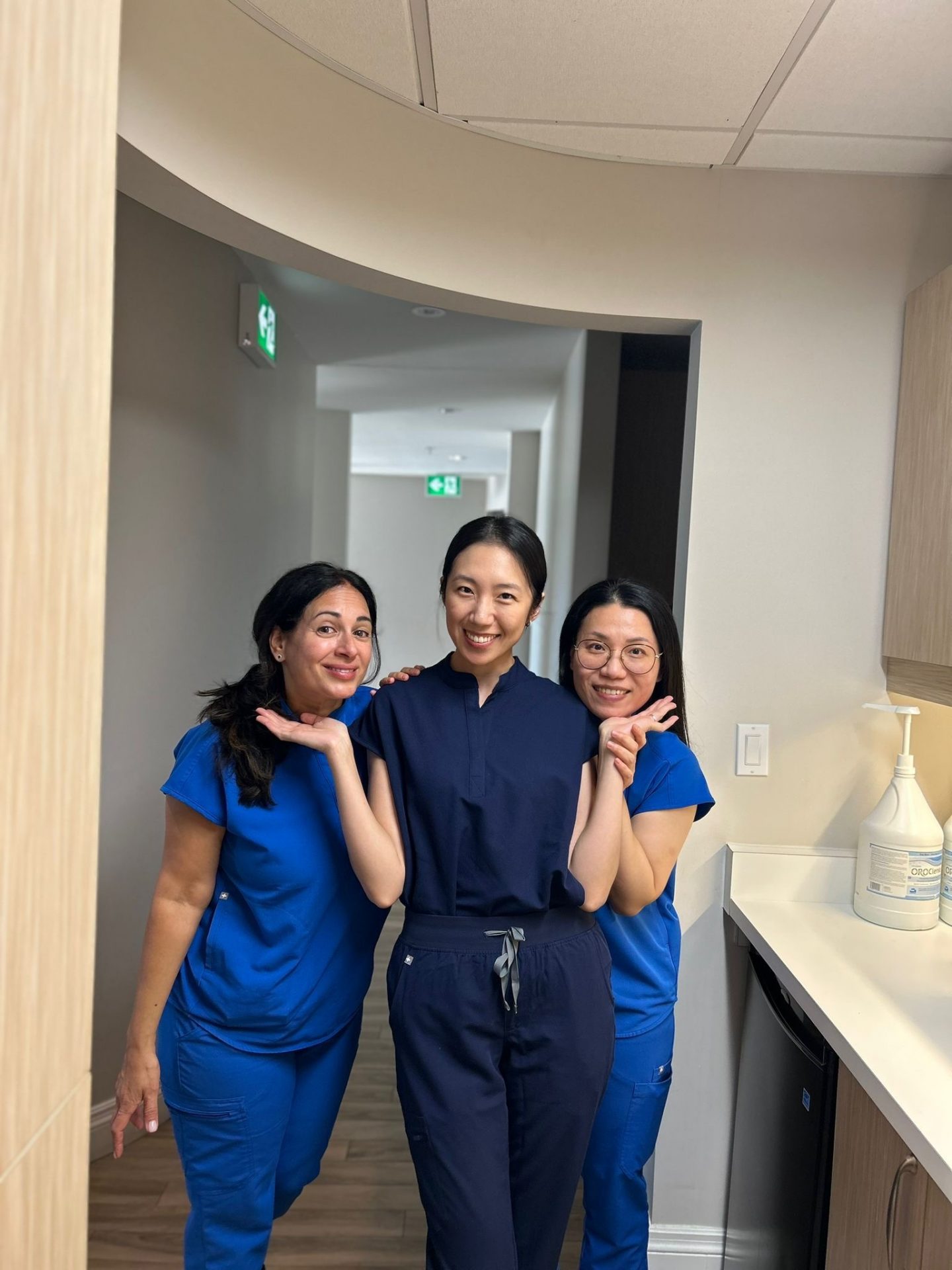 Three people in blue scrubs smiling, posing in a hallway. They stand close together, brightening the neutral-toned interior space.