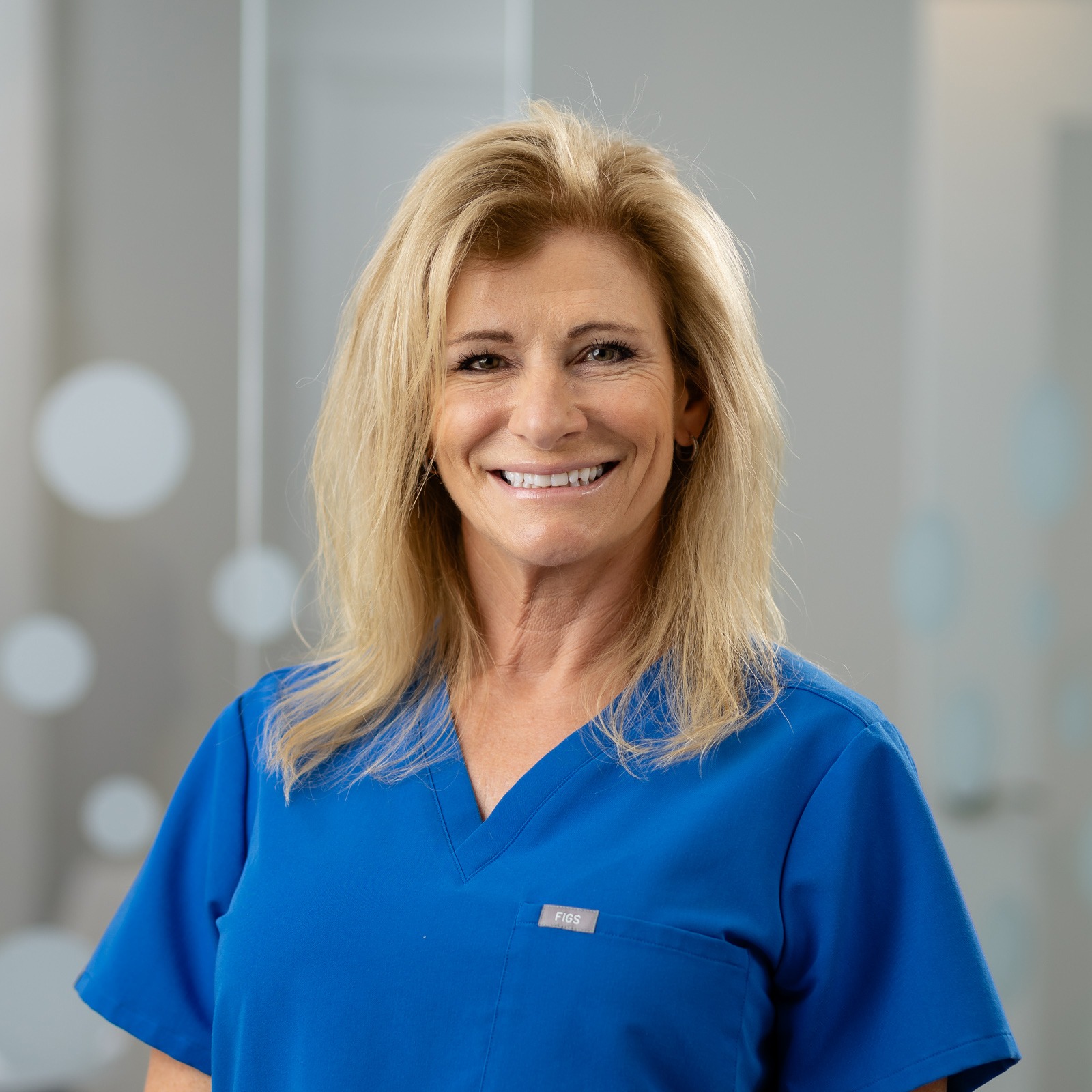 A person in a blue scrub top smiles in a bright room, featuring frosted glass with circle patterns in the background.