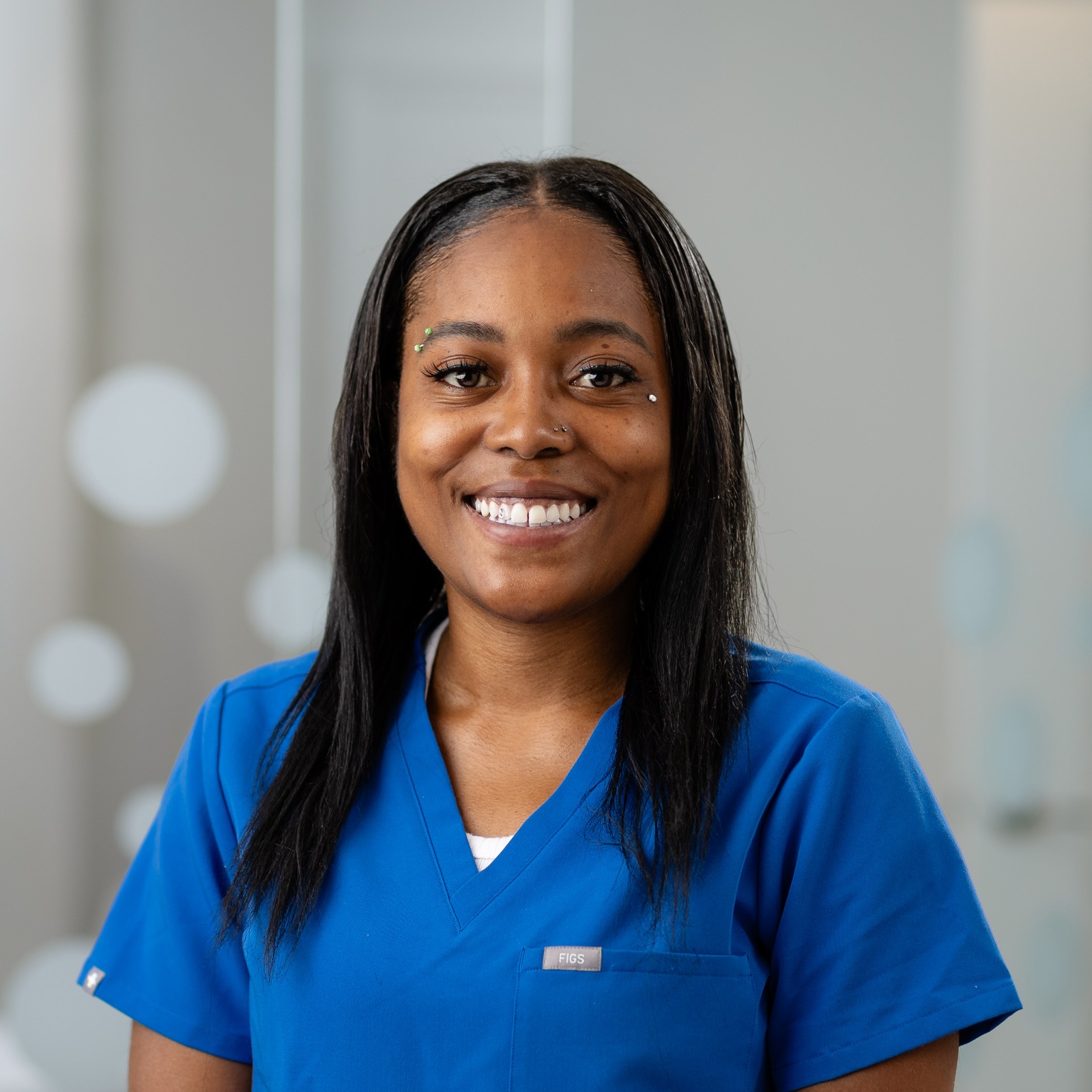 A person wearing blue scrubs smiles warmly, standing indoors with a blurred background and circular light reflections.