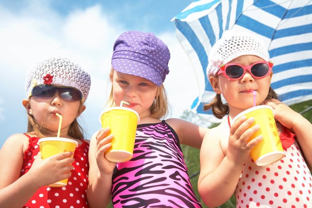 Three children in swimsuits and hats drinking from yellow cups under a blue and white umbrella on a sunny day.