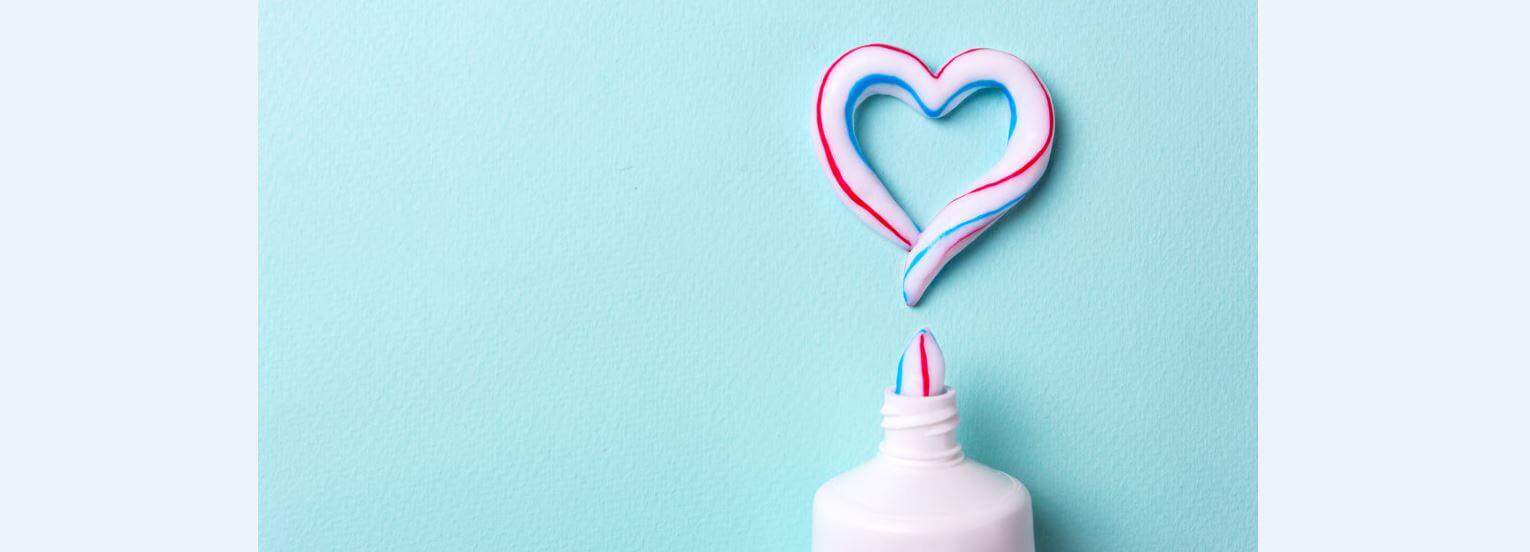A toothpaste tube squirts red, white, blue stripes forming a heart shape against a light blue background, symbolizing freshness and care.
