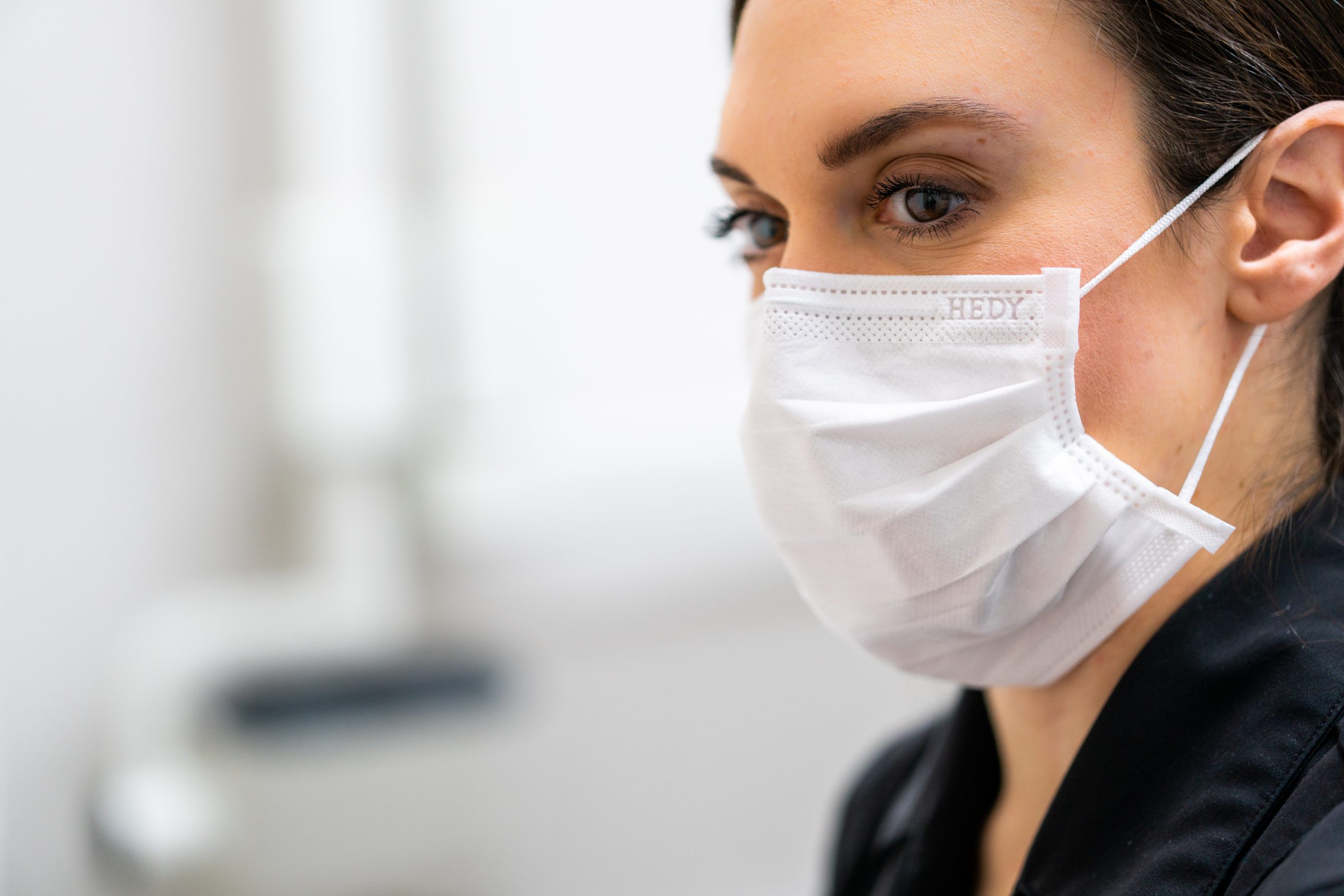 A person wearing a white surgical mask with "HEDY" printed on it, focusing intently. Background is out of focus.