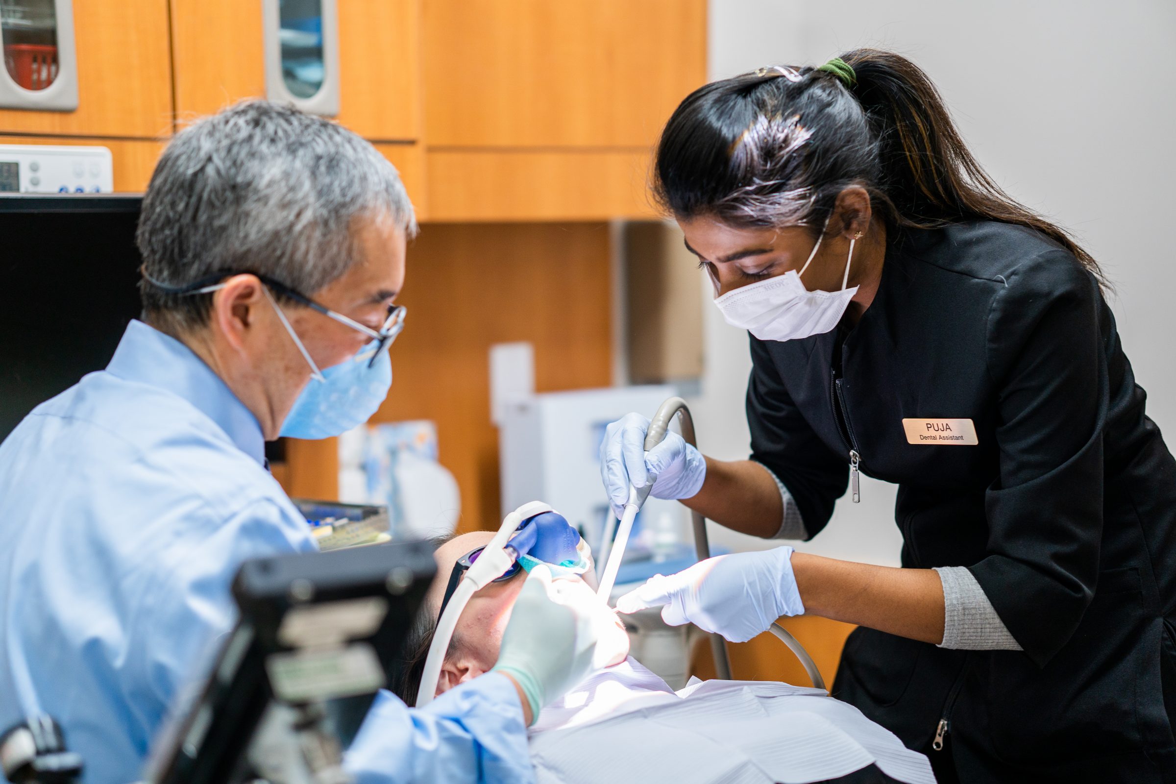 Two dental professionals perform a procedure on a patient in a clinic, wearing masks and gloves, focused on oral care.