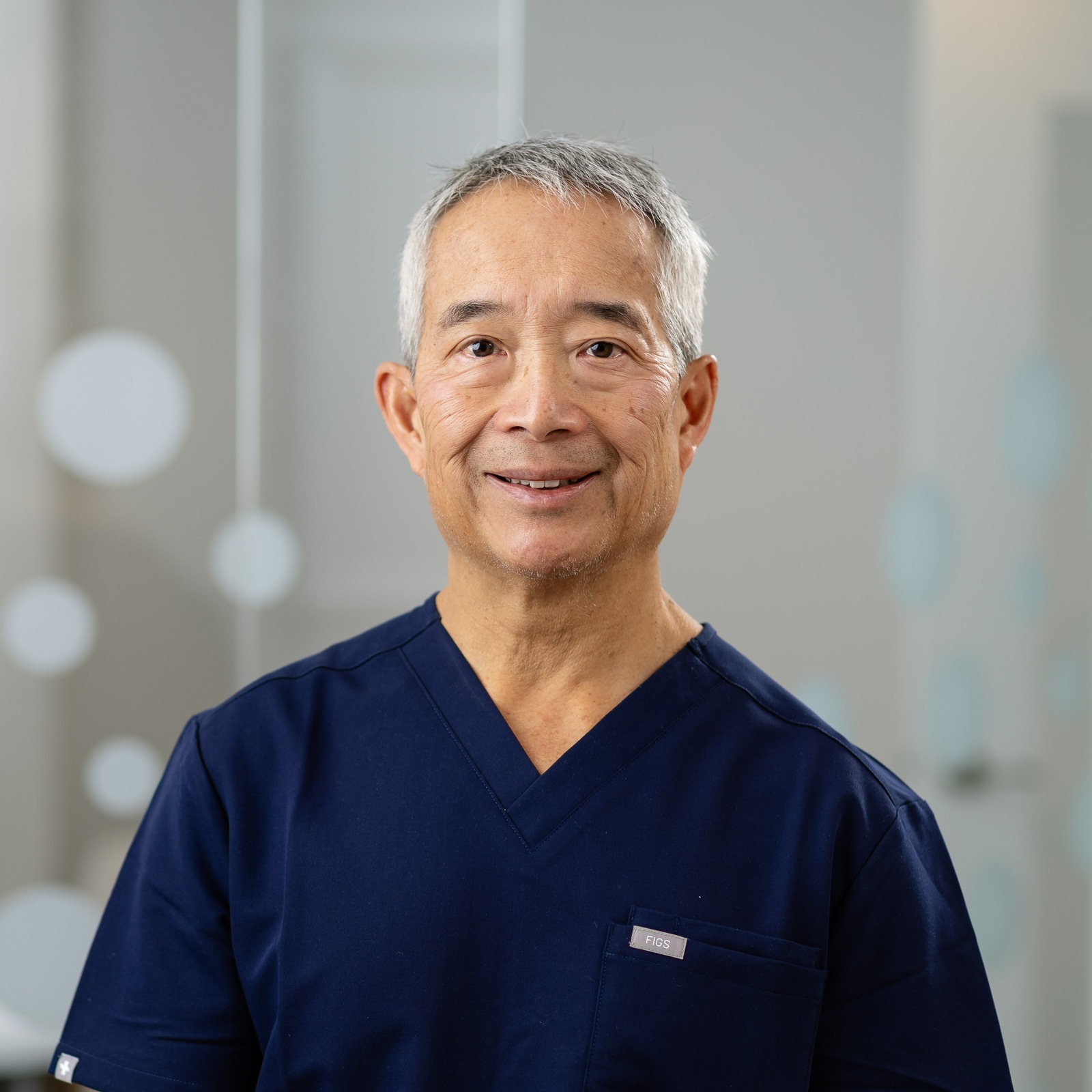 A smiling person in a blue medical uniform stands indoors, with a blurred, light background featuring circular patterns on a glass surface.