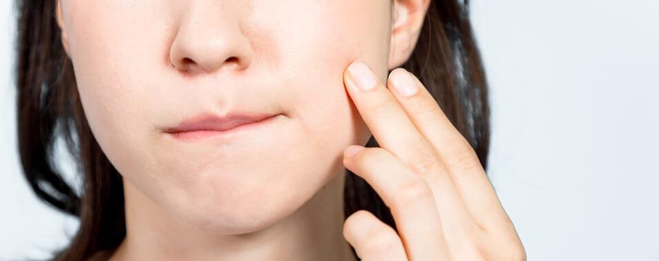 A person touches their cheek with fingertips, lips pressed together, conveying contemplation or concern against a plain background.