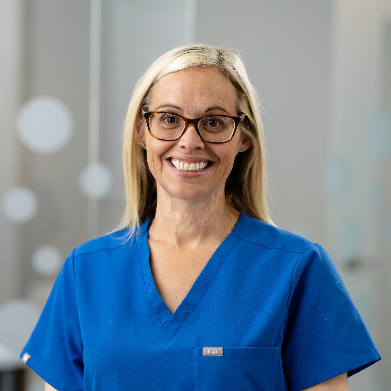 A person wearing blue medical scrubs smiles while standing indoors. The background is blurred with circular light spots, suggesting a clinical environment.