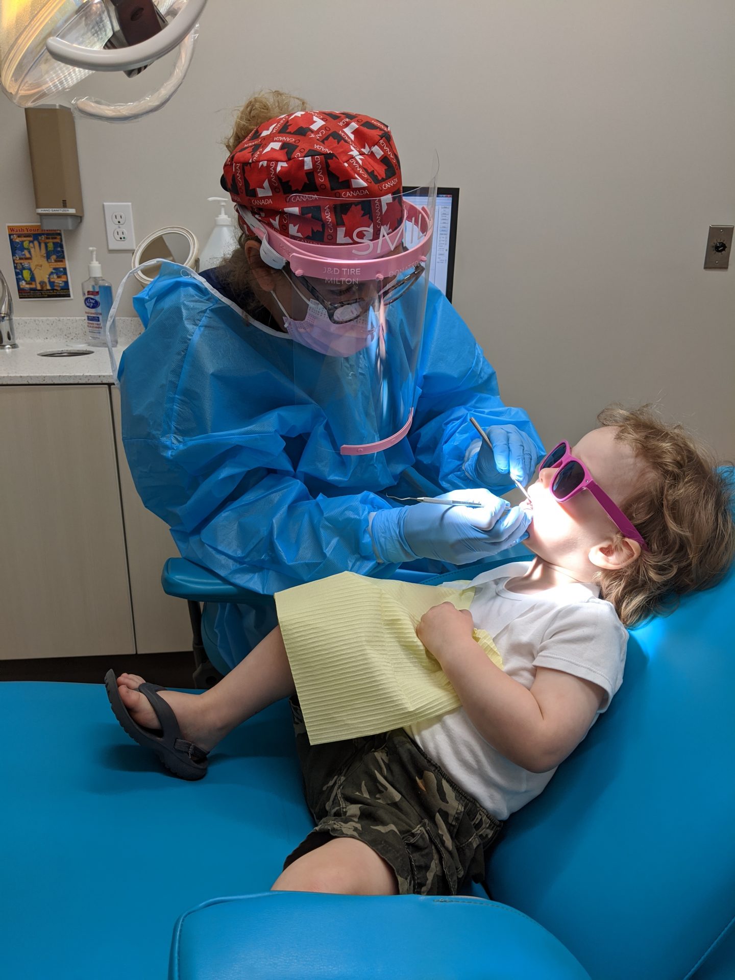 A person in protective gear examines a child's teeth in a brightly-lit dental office, with the child wearing sunglasses and a bib.