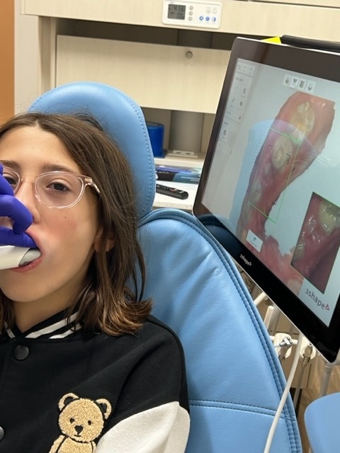 A person is sitting in a dental chair with a tool in their mouth, while a screen displays dental images.