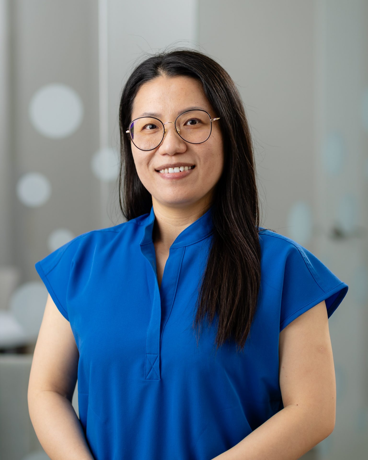 A person in a blue shirt smiles indoors, wearing glasses, with a soft, neutral background featuring circular patterns.