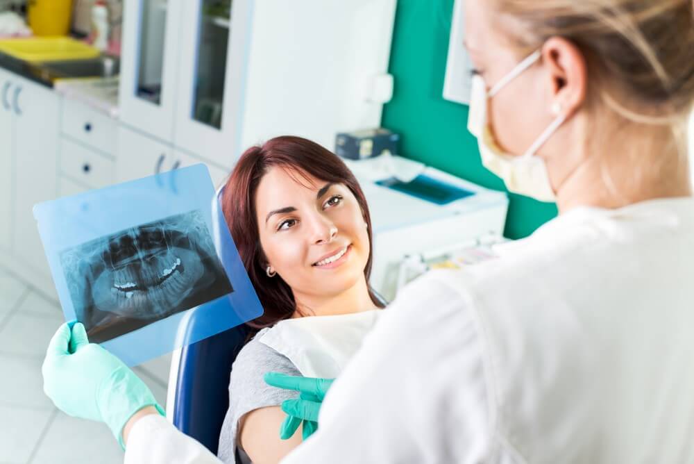 Two people in a dental office, one holding an X-ray image. The other is seated, smiling, and wearing a dental bib.