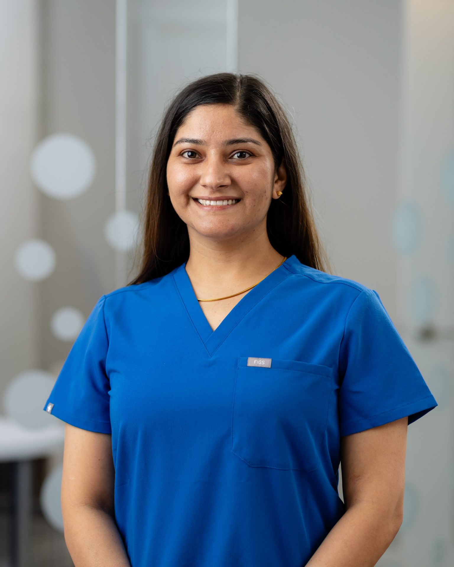 A person wearing a blue medical scrub top smiles indoors, standing against a softly blurred, light-colored background with circular patterns.