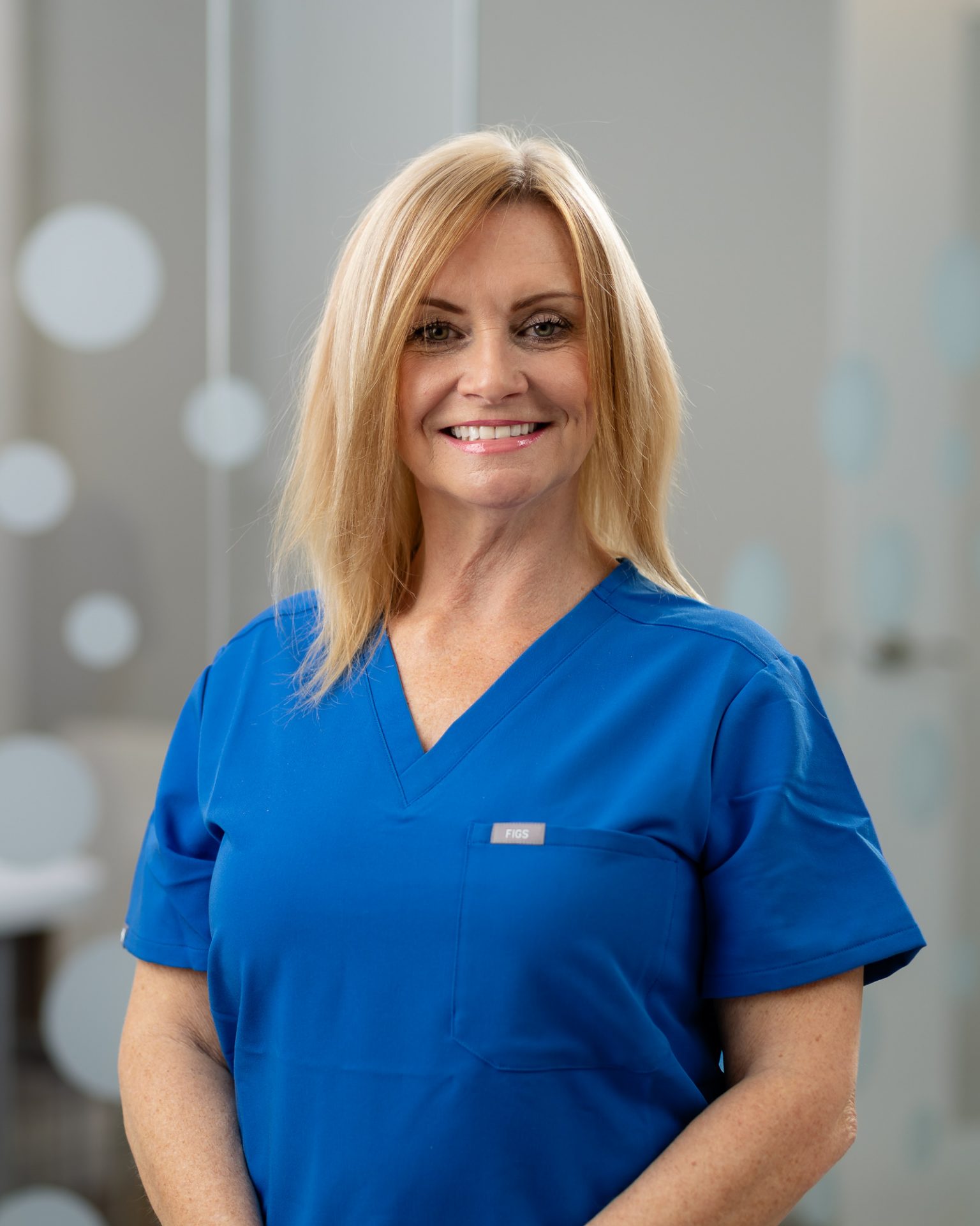 A person wearing blue medical scrubs stands indoors, smiling. The background features a modern design with circular patterns on glass panels.