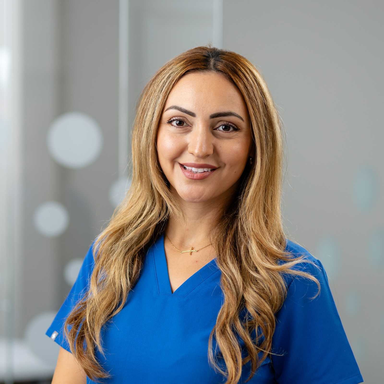 A person wearing blue scrubs smiles in a modern setting with glass partitions, decorated with circular patterns, and soft gray background.