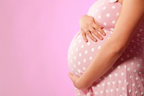 A person in a pink polka dot dress gently cradles a pregnant belly against a plain pink background, exuding warmth and anticipation.