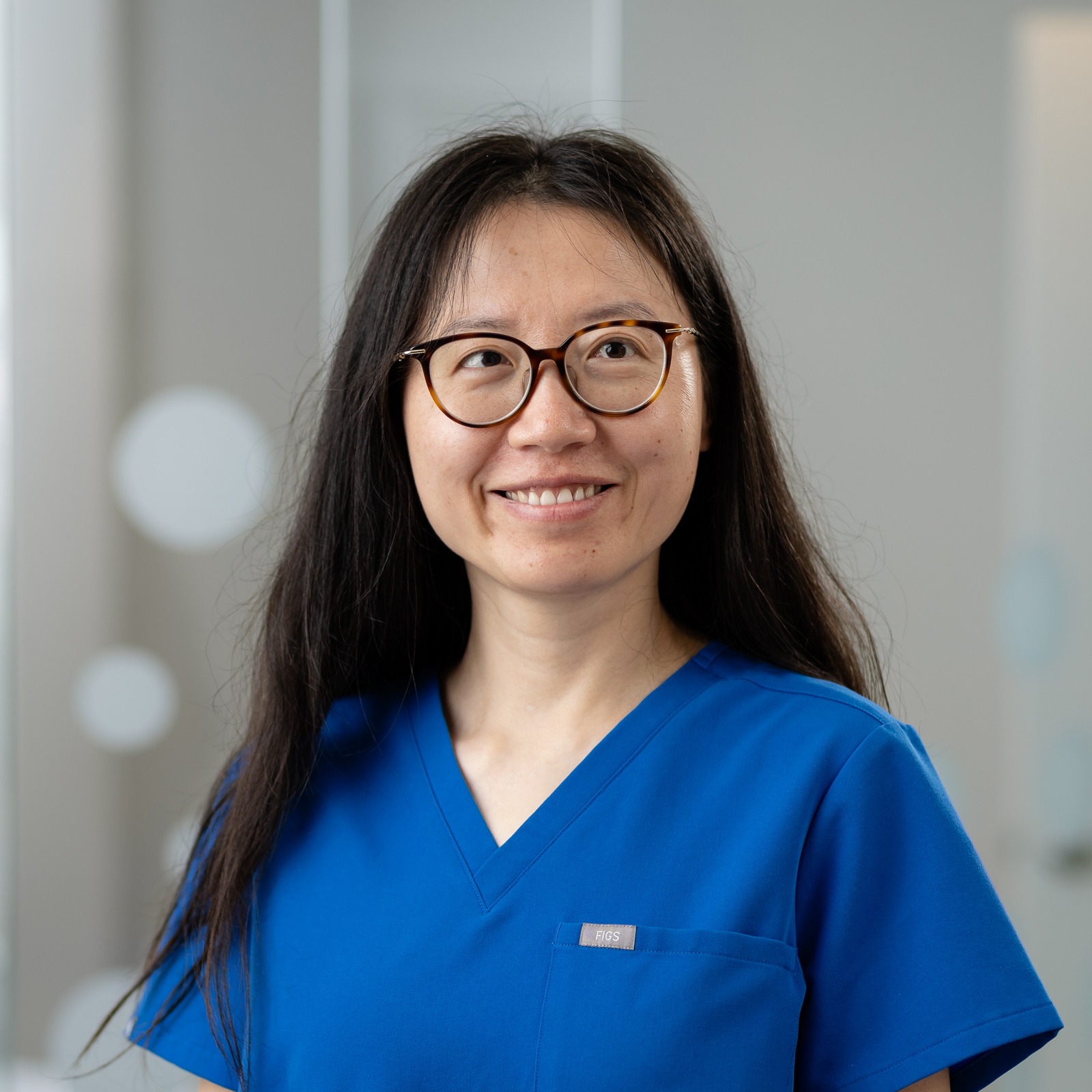 A person with long hair and glasses wearing blue medical scrubs smiles in a bright, modern room.