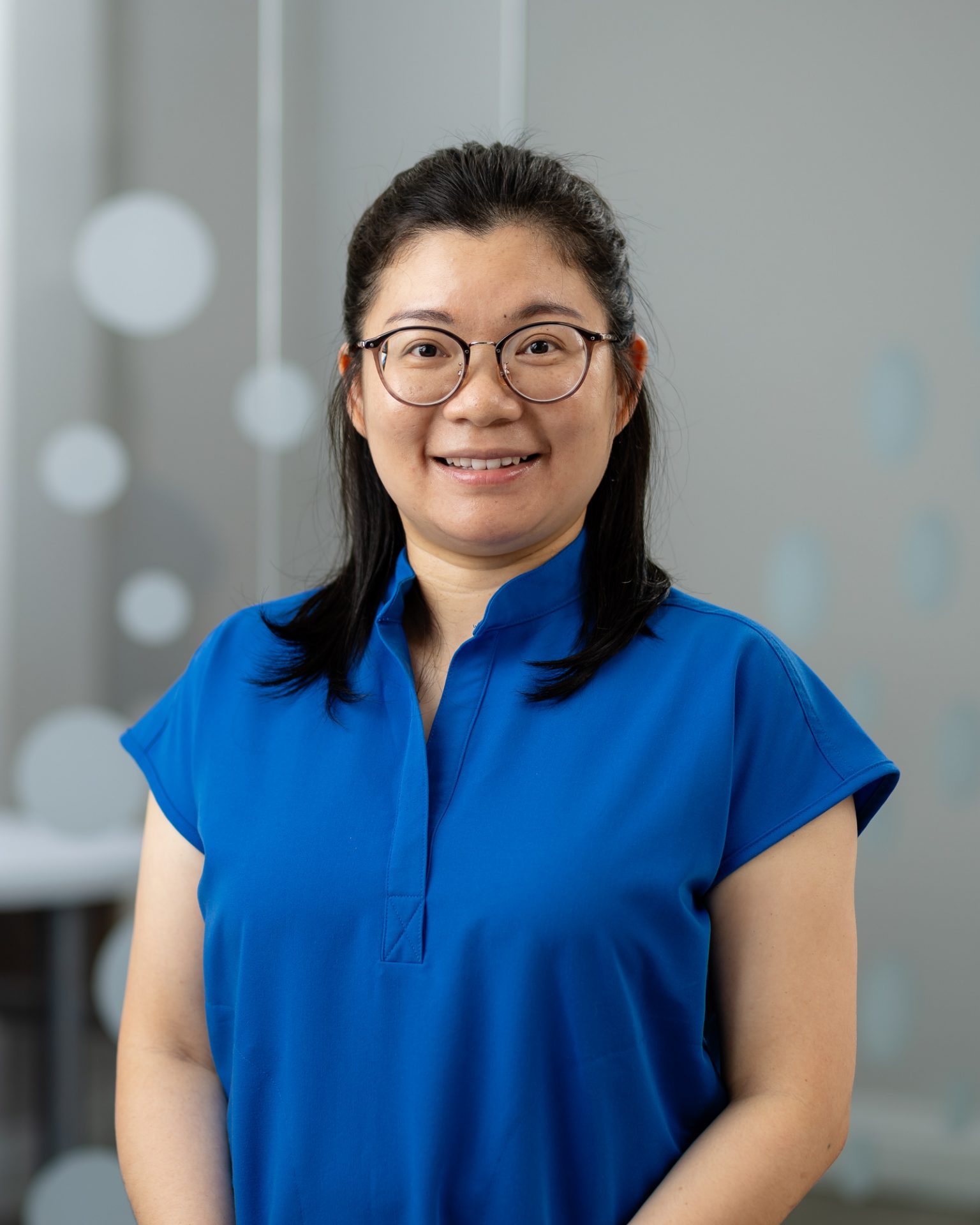 A person wearing glasses and a blue shirt smiles in a studio setting with a blurred, modern background featuring circular decorations.