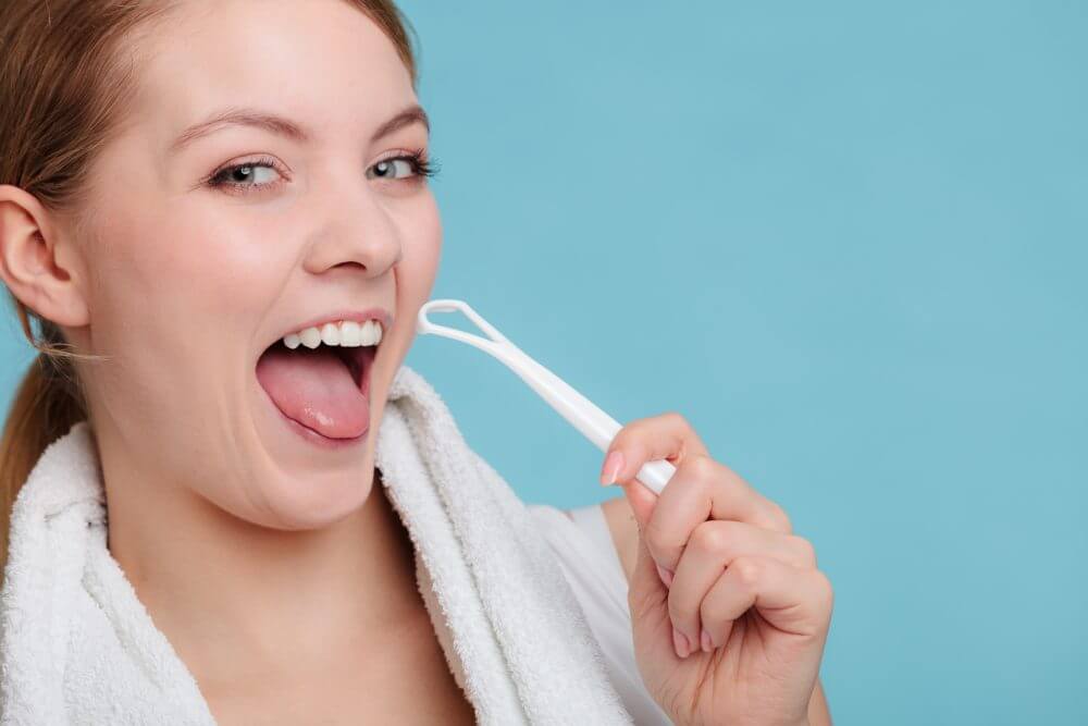 A person with a towel around their neck uses a tongue scraper, smiling against a plain blue background.