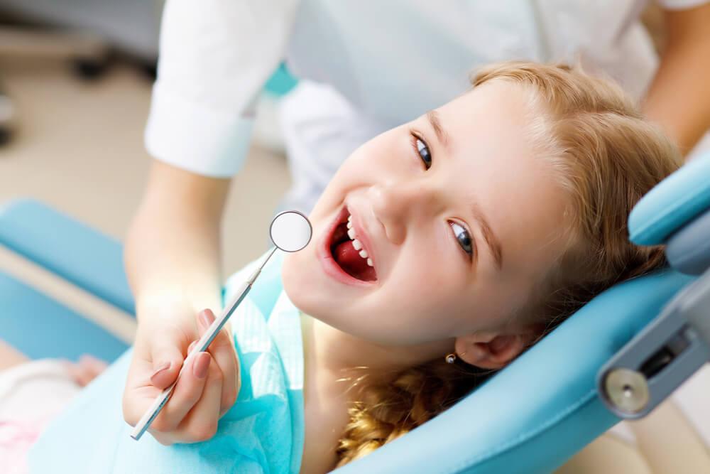 A child wearing a blue bib smiles in a dental chair, holding a dentist's mirror, with a person nearby in a white coat.