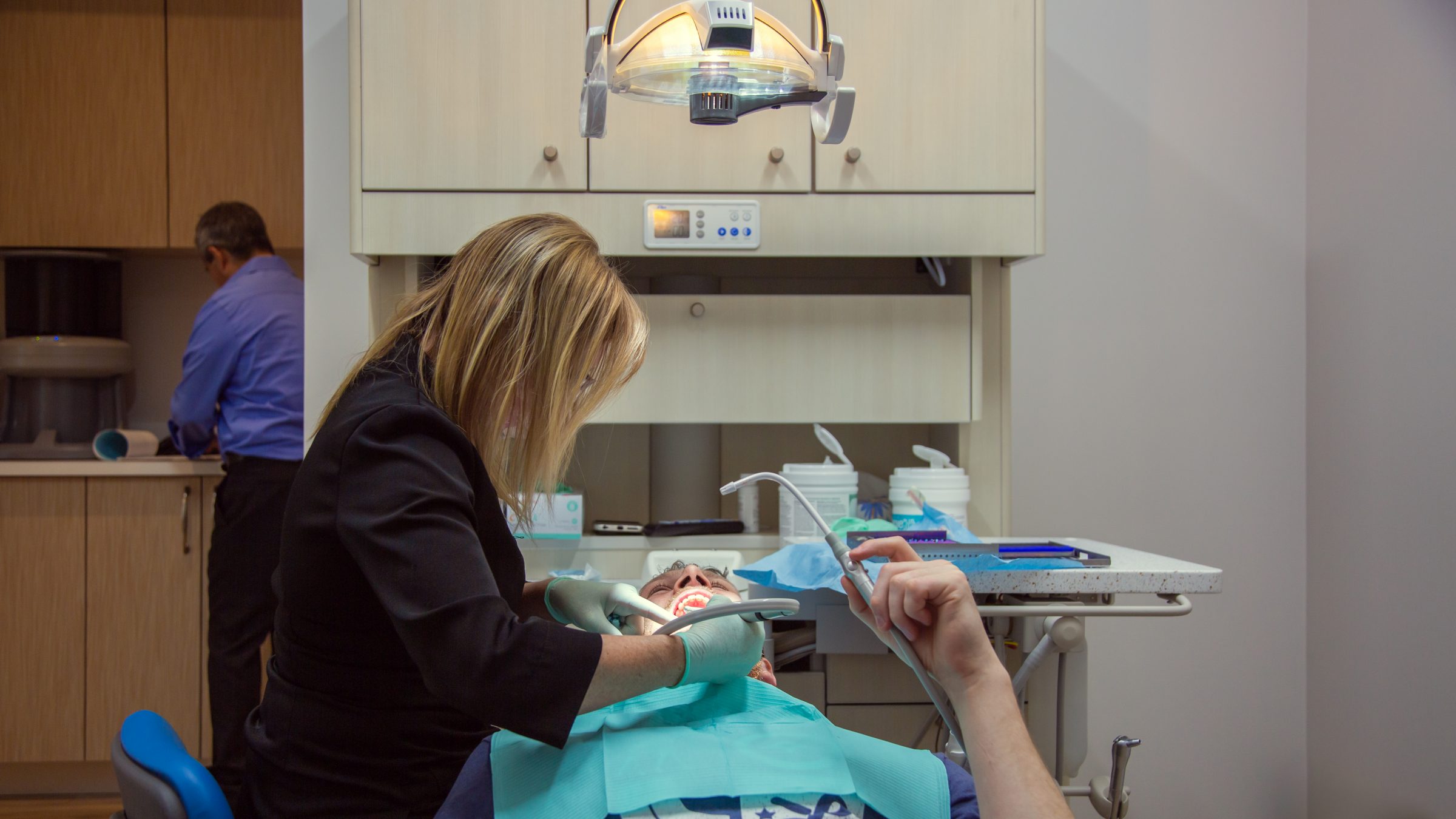 A person receives dental treatment under a bright light, while another person stands in the background near cabinets and equipment.