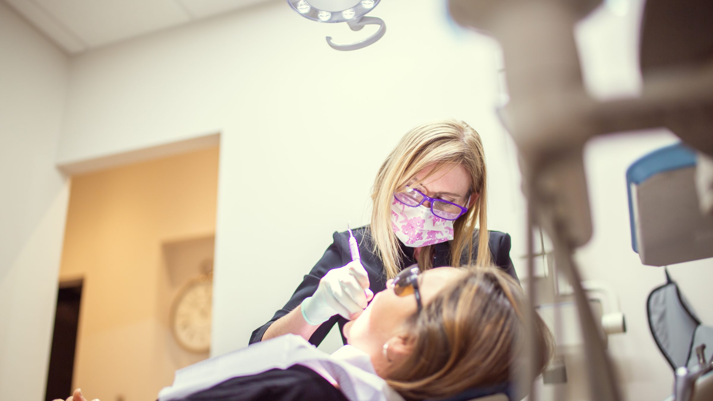 A person wearing a mask and glasses performs a dental procedure on another person in a brightly lit clinic room.