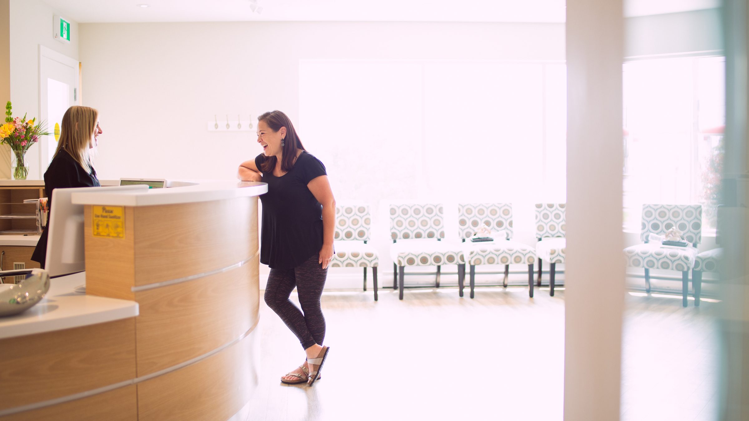 Two people interact at a bright, modern reception desk. The waiting area features patterned chairs and flowers enhance the welcoming atmosphere.