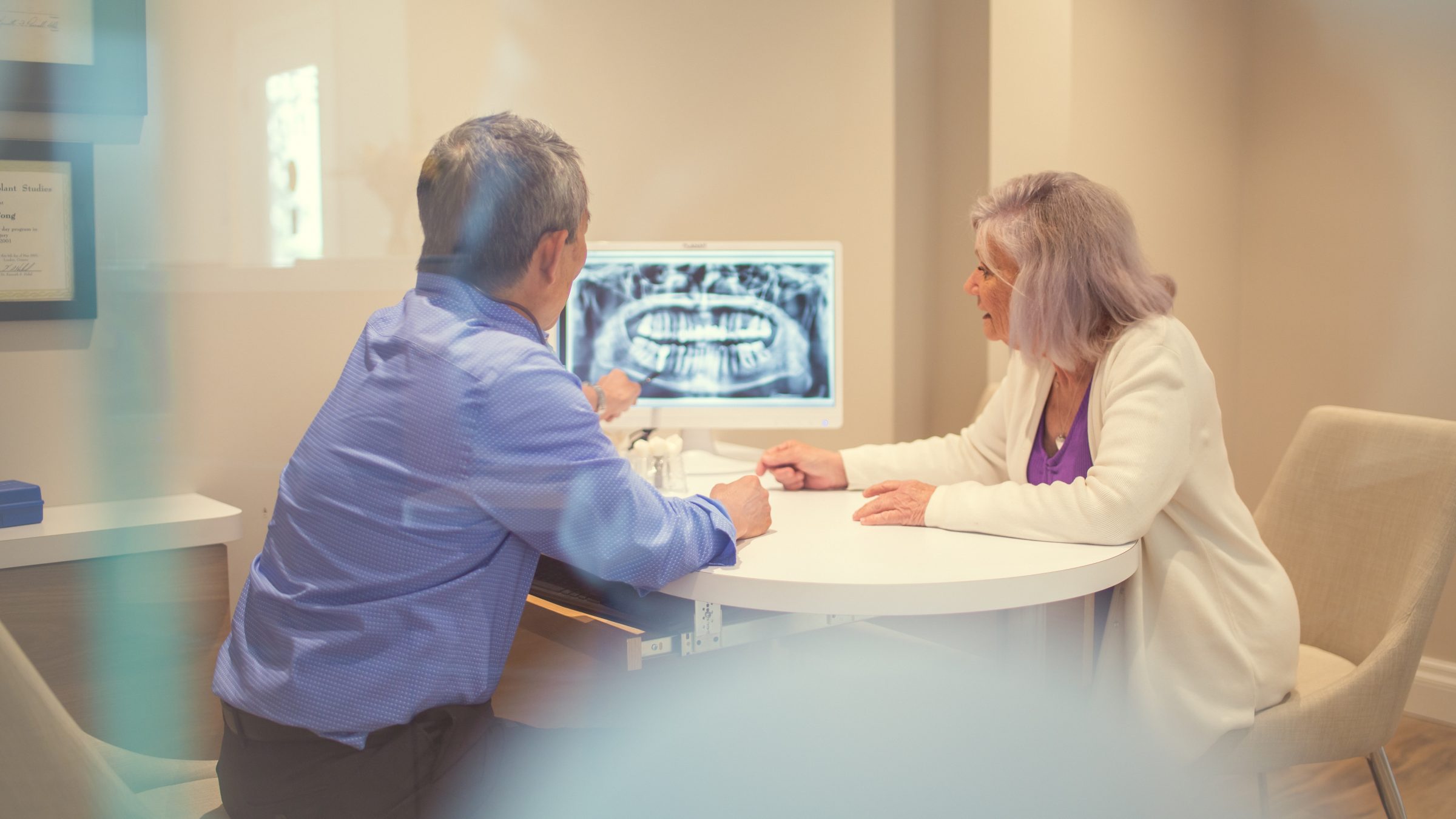 Two people in an office setting discuss dental X-rays displayed on a computer screen, seated around a small table.