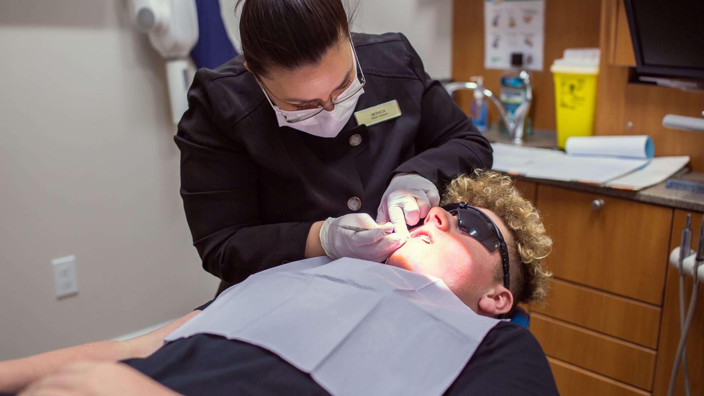 A dental professional examines a patient's teeth in a clinic. The patient wears sunglasses, and dental tools are visible.