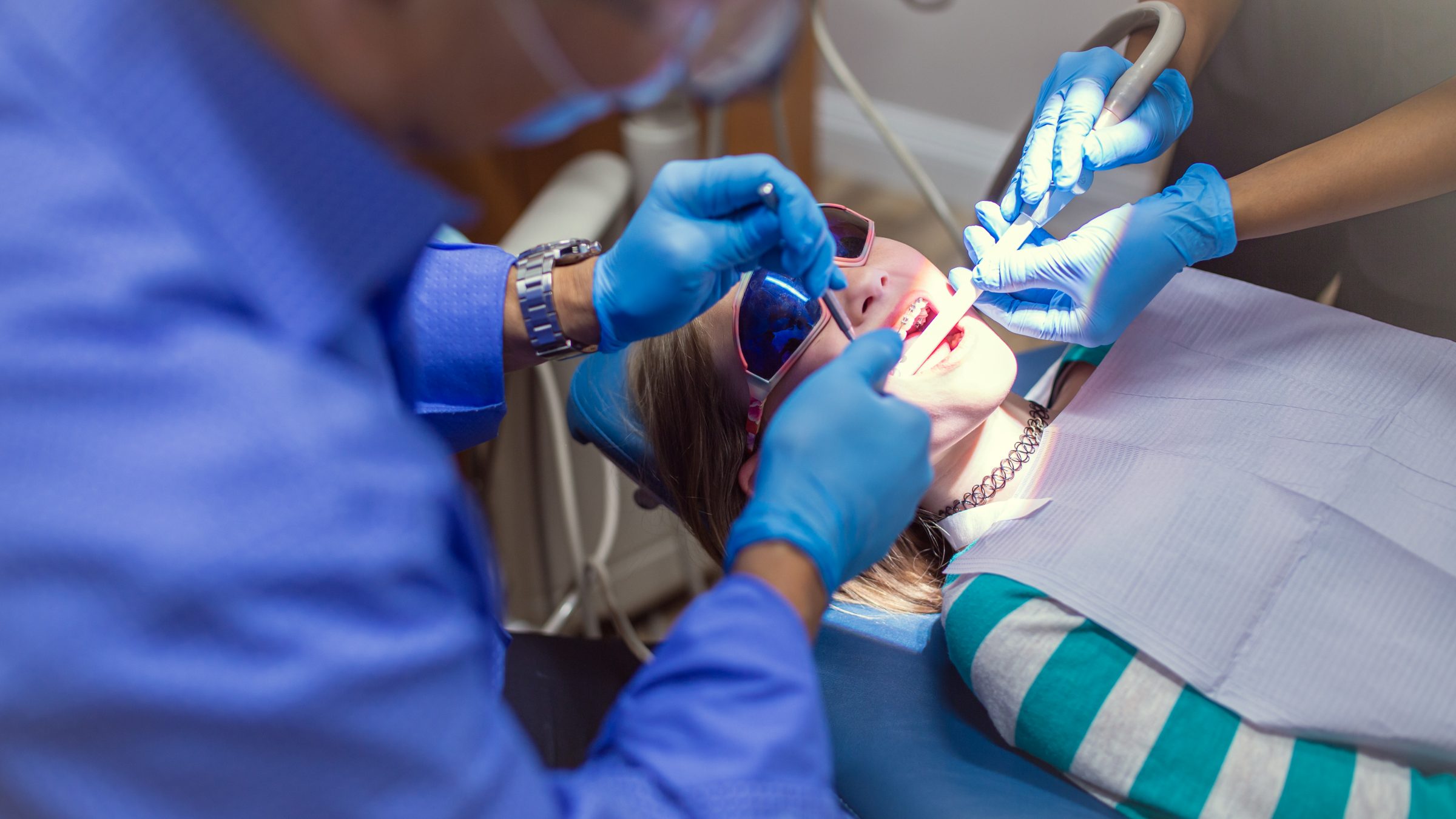 A person is focused on dental work, examining a patient's teeth. The patient wears sunglasses, and both individuals have protective gloves on.