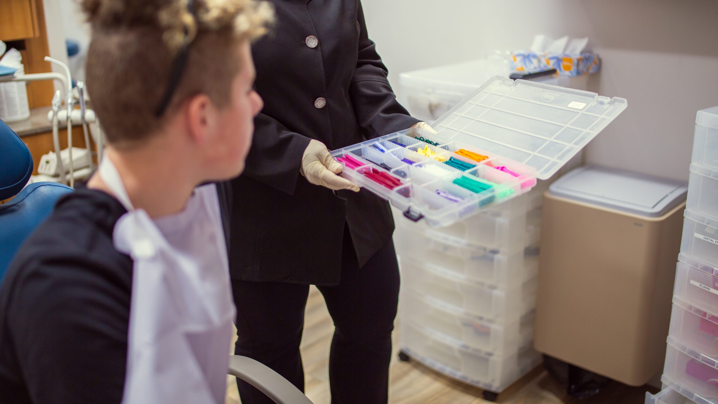 A person in a dental setting examines a colorful box of dental tools while another person sits nearby, wearing a protective bib.