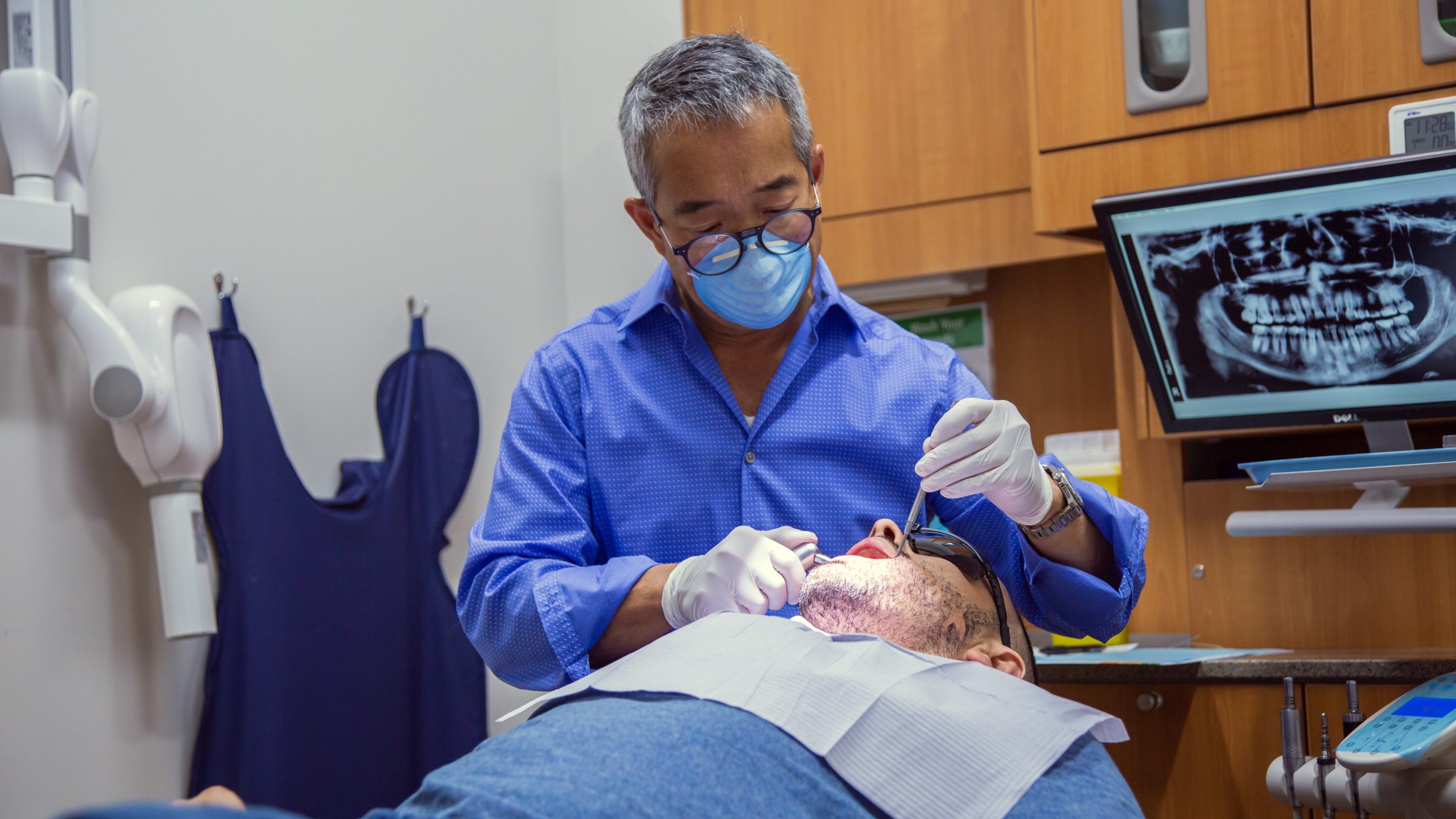 A person wearing a blue shirt and mask examines another person’s teeth in a dental office with an X-ray display and dental tools.