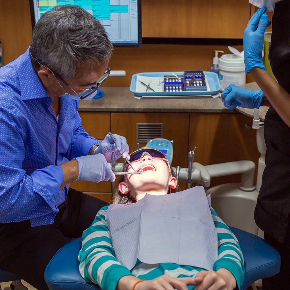 A dentist examines a child's teeth in a modern dental office, with instruments and a computer screen in the background.