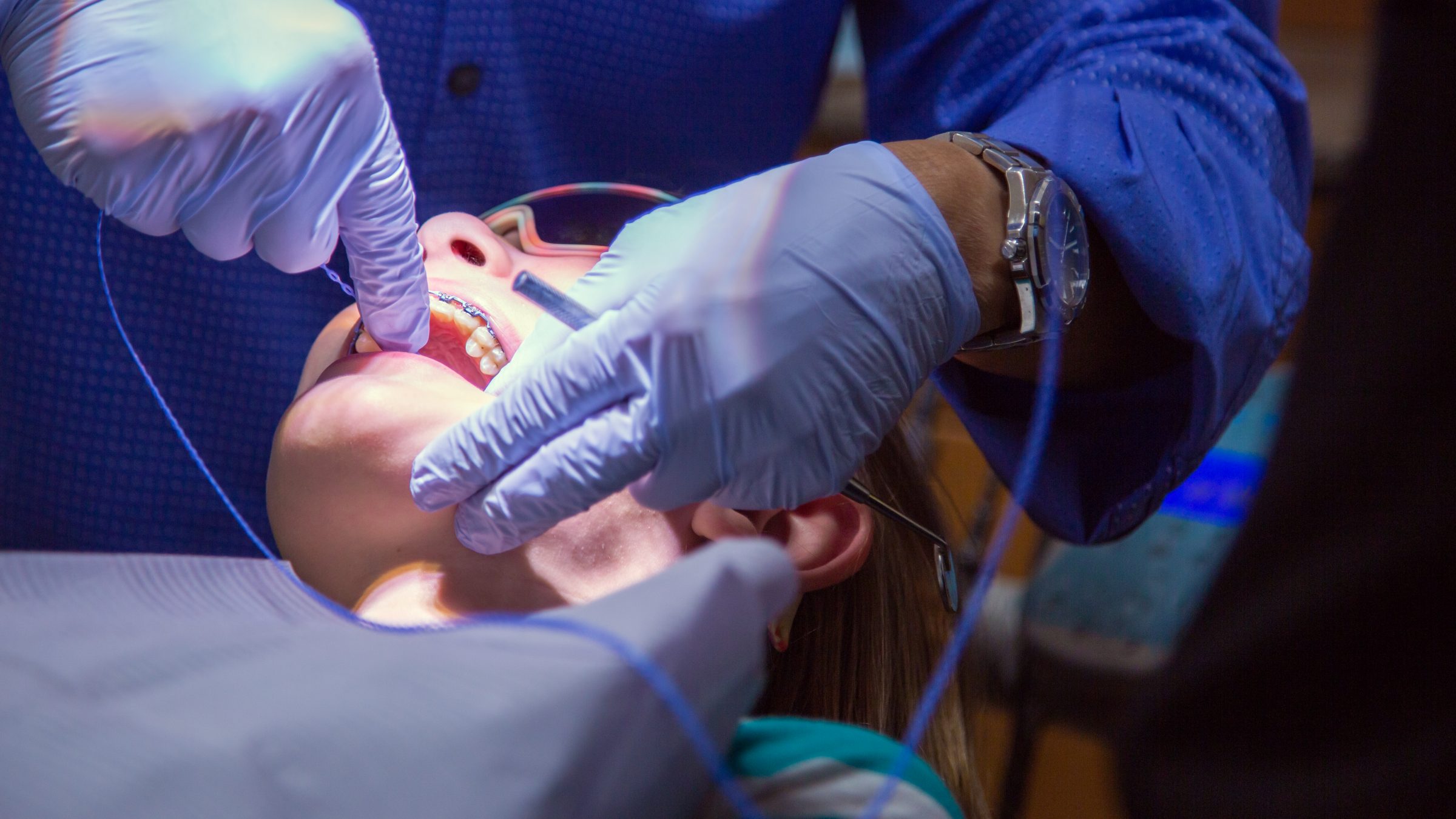 A person wearing gloves examines a patient's teeth with braces under bright lighting in a dental clinic setting.