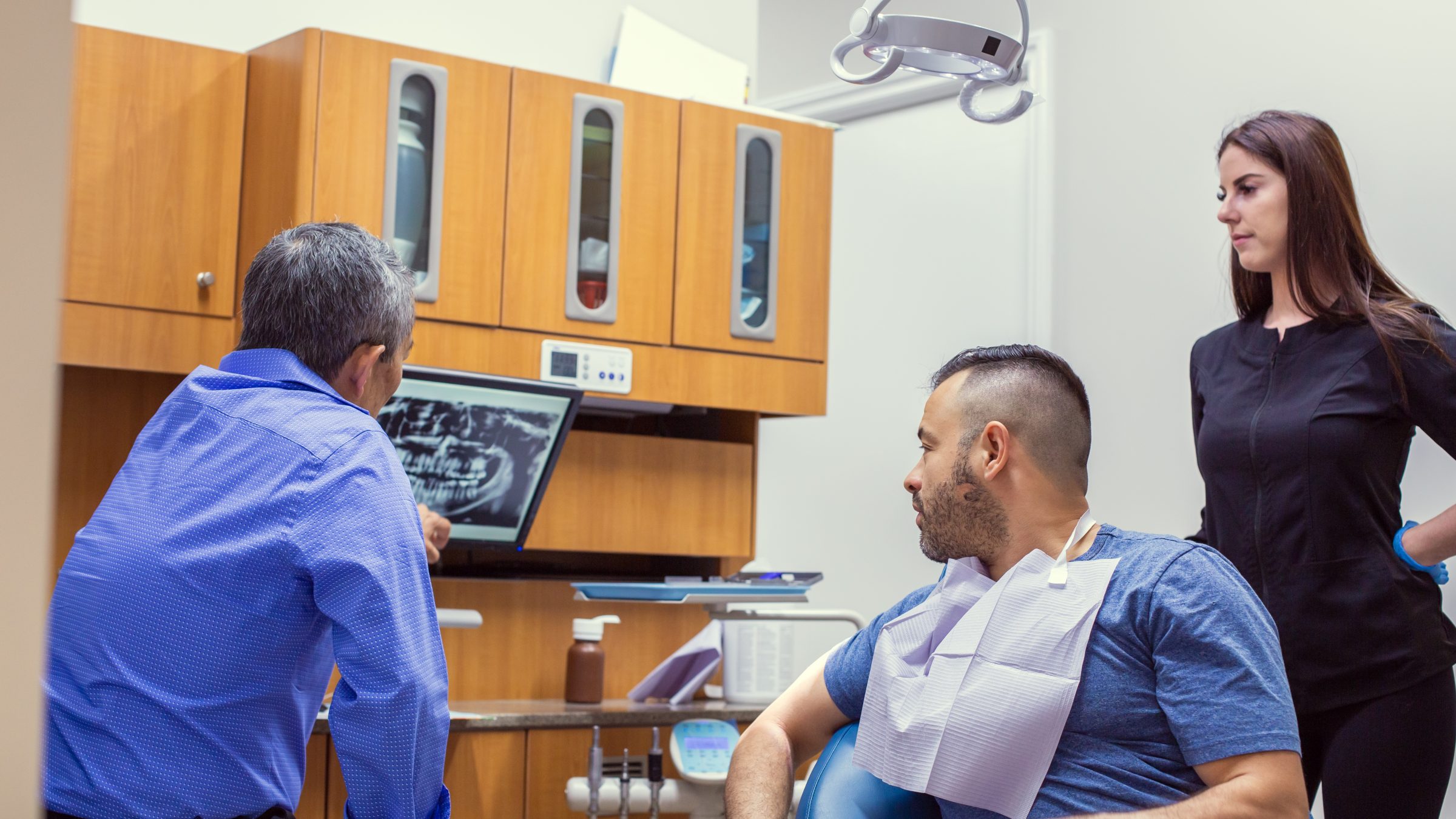 A person in a dental office showing an X-ray to a seated patient, with another person observing, surrounded by wooden cabinets.