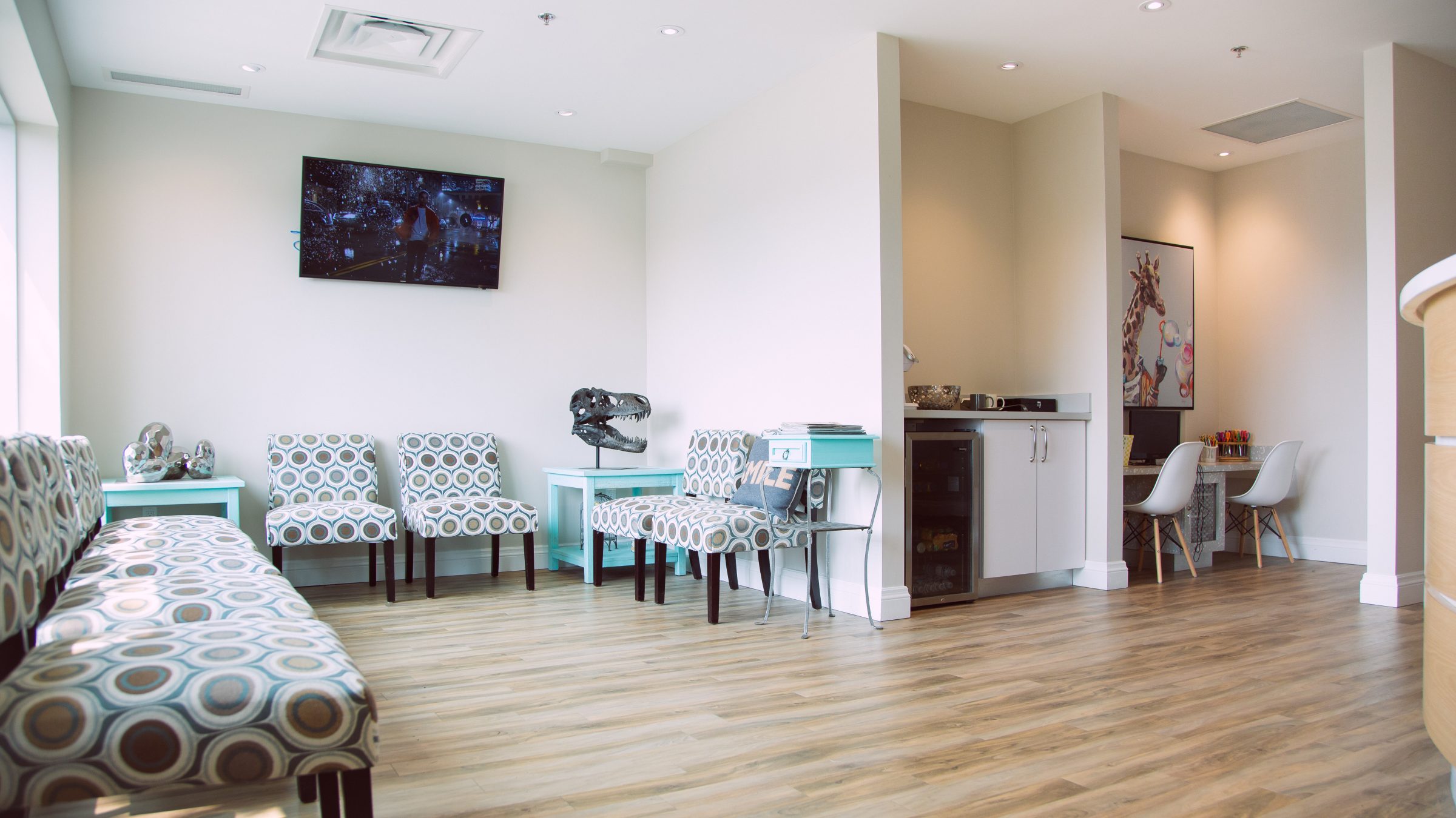 Modern waiting room with patterned chairs, TV, dinosaur sculpture, and art featuring a giraffe blowing bubbles. Light wood floors and natural lighting.