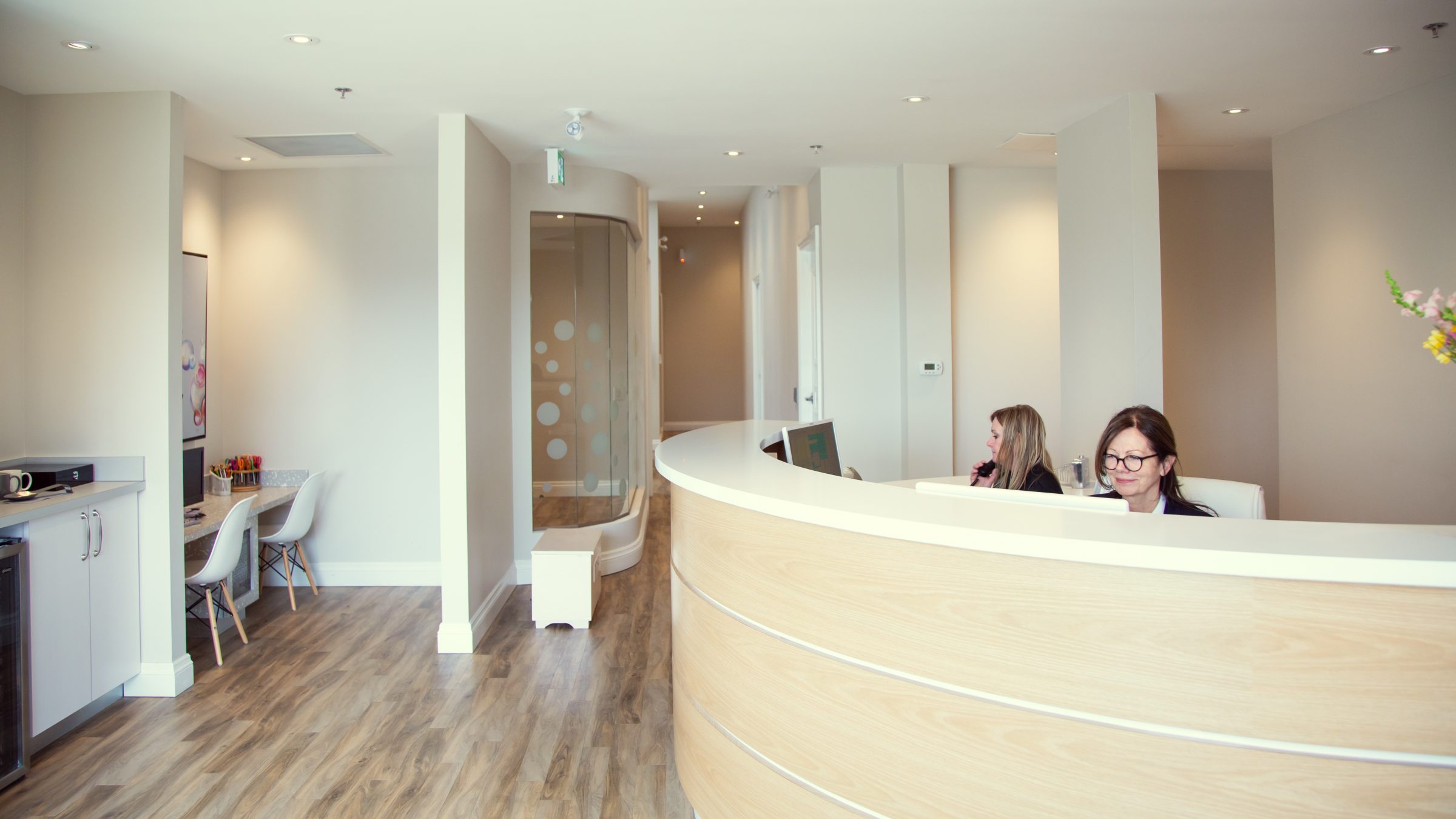 Modern dental office with two receptionists at a curved desk. Light wood flooring and minimalist decor create a welcoming and clean atmosphere.