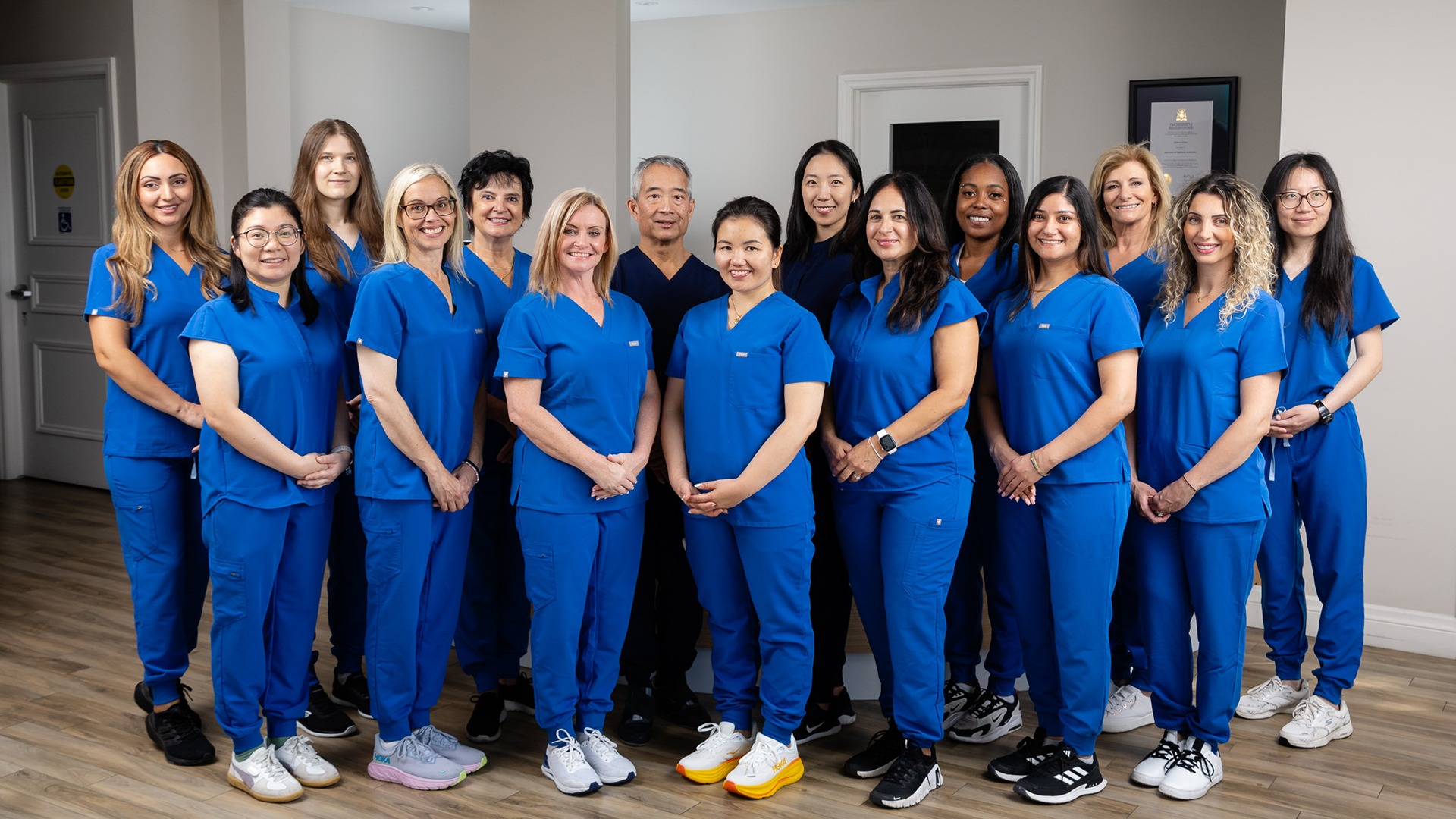 Fifteen people in blue uniforms stand indoors on a wooden floor, facing forward and smiling, with neutral-colored walls in the background.