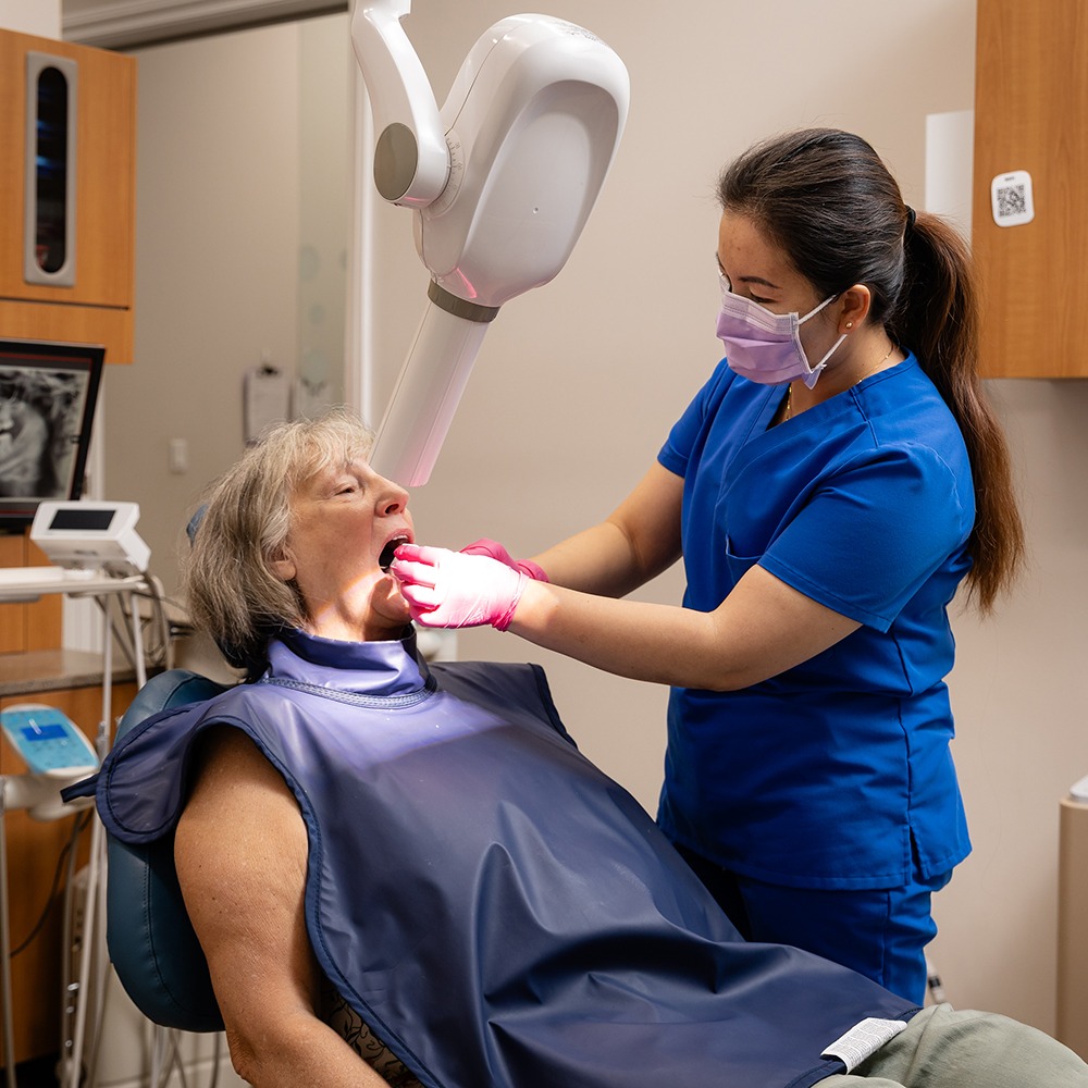 A person in blue scrubs examines another person's teeth in a dental office. Equipment and wooden cabinets are visible in the background.