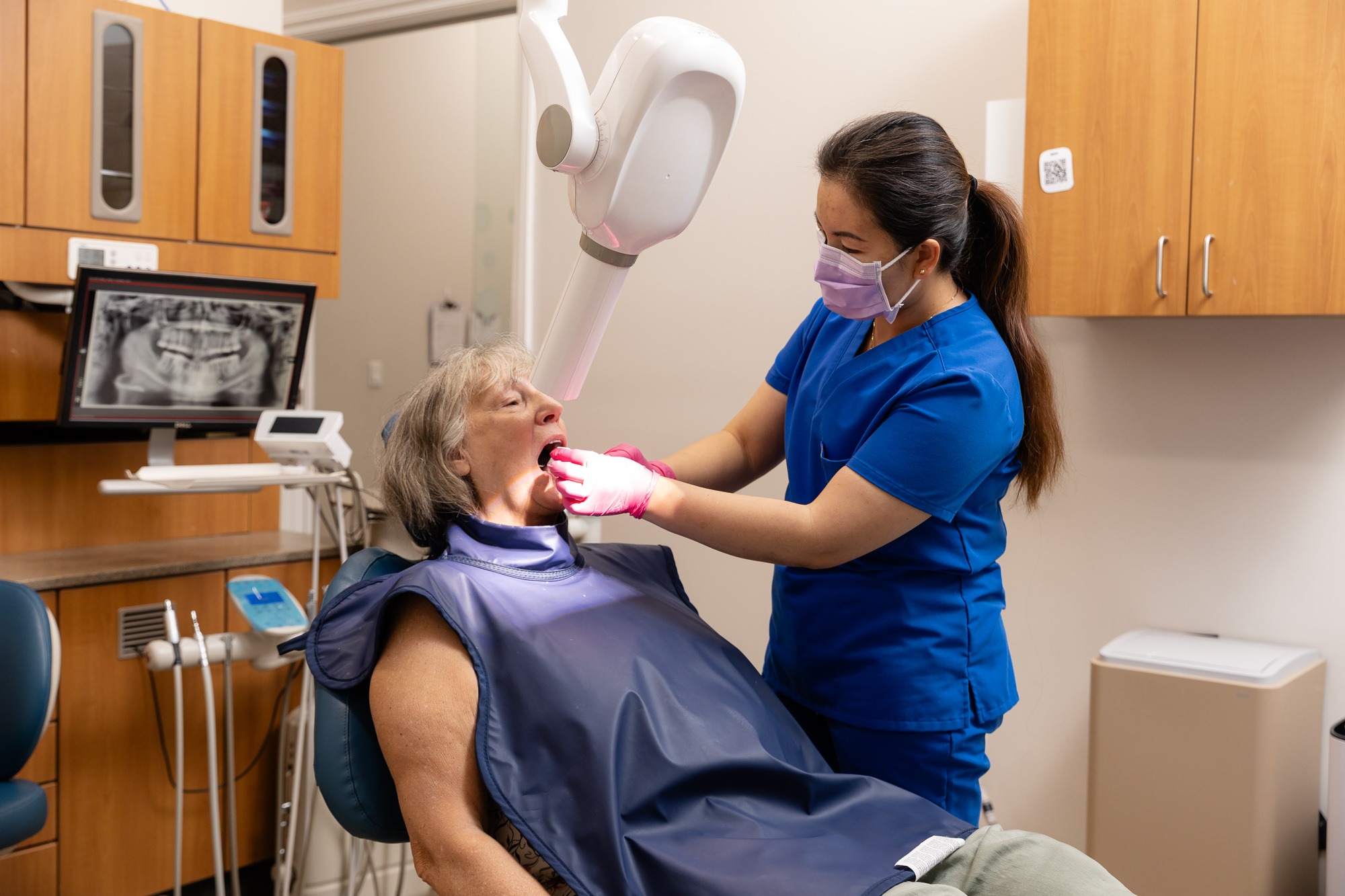 A dental professional in blue scrubs examines a person's mouth in a clinic, with dental tools and an X-ray monitor.