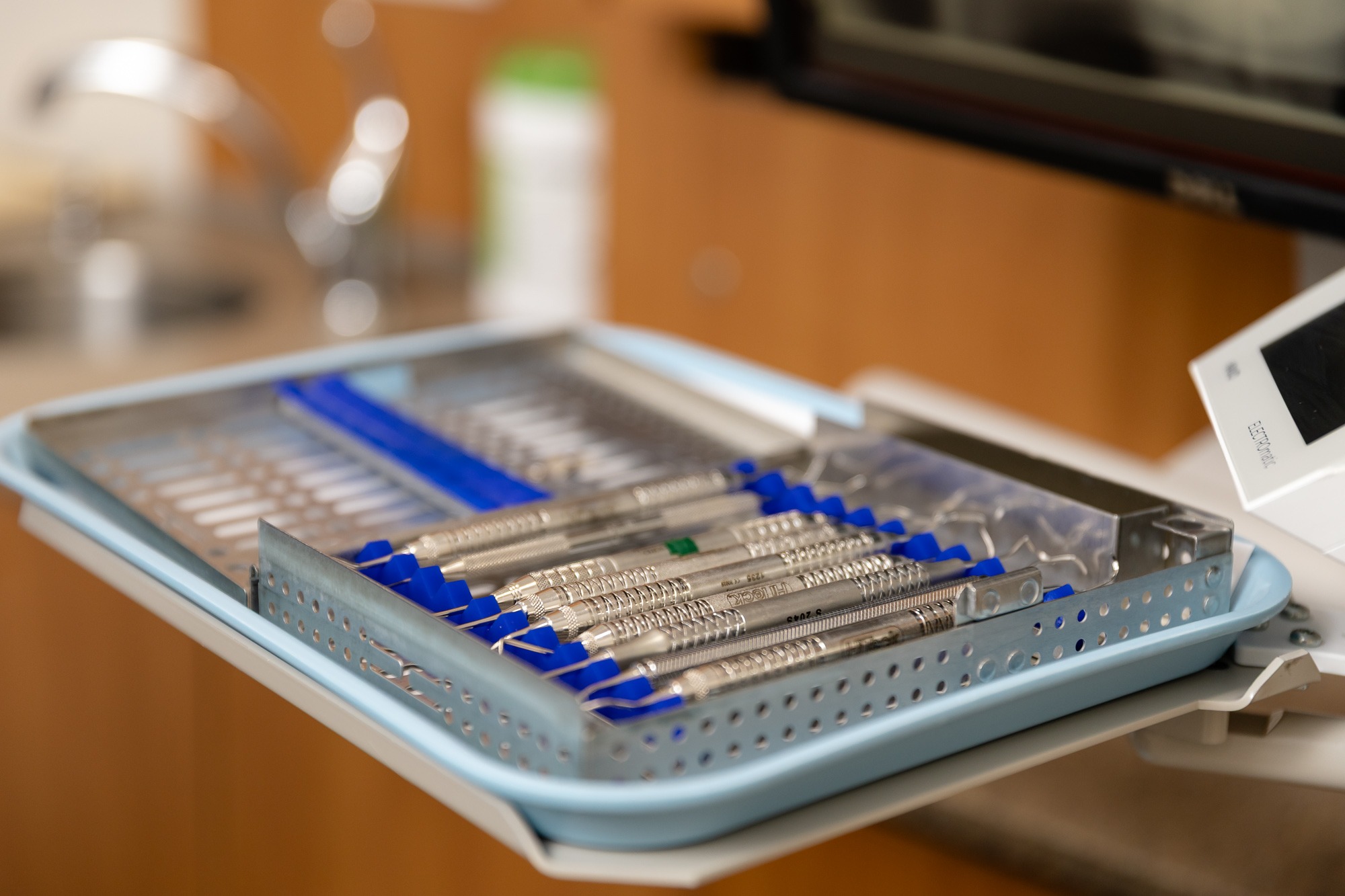 A dental setup with sterilized tools in a blue tray, located in a clinic setting with a blurred sink background.