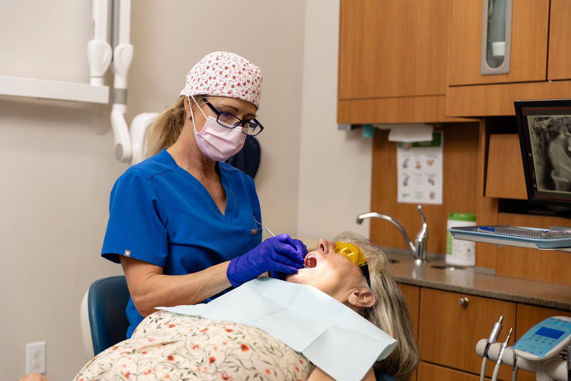 A dental professional in scrubs and mask examines a patient in a dental chair inside a clinic, using tools and wearing protective eyewear.