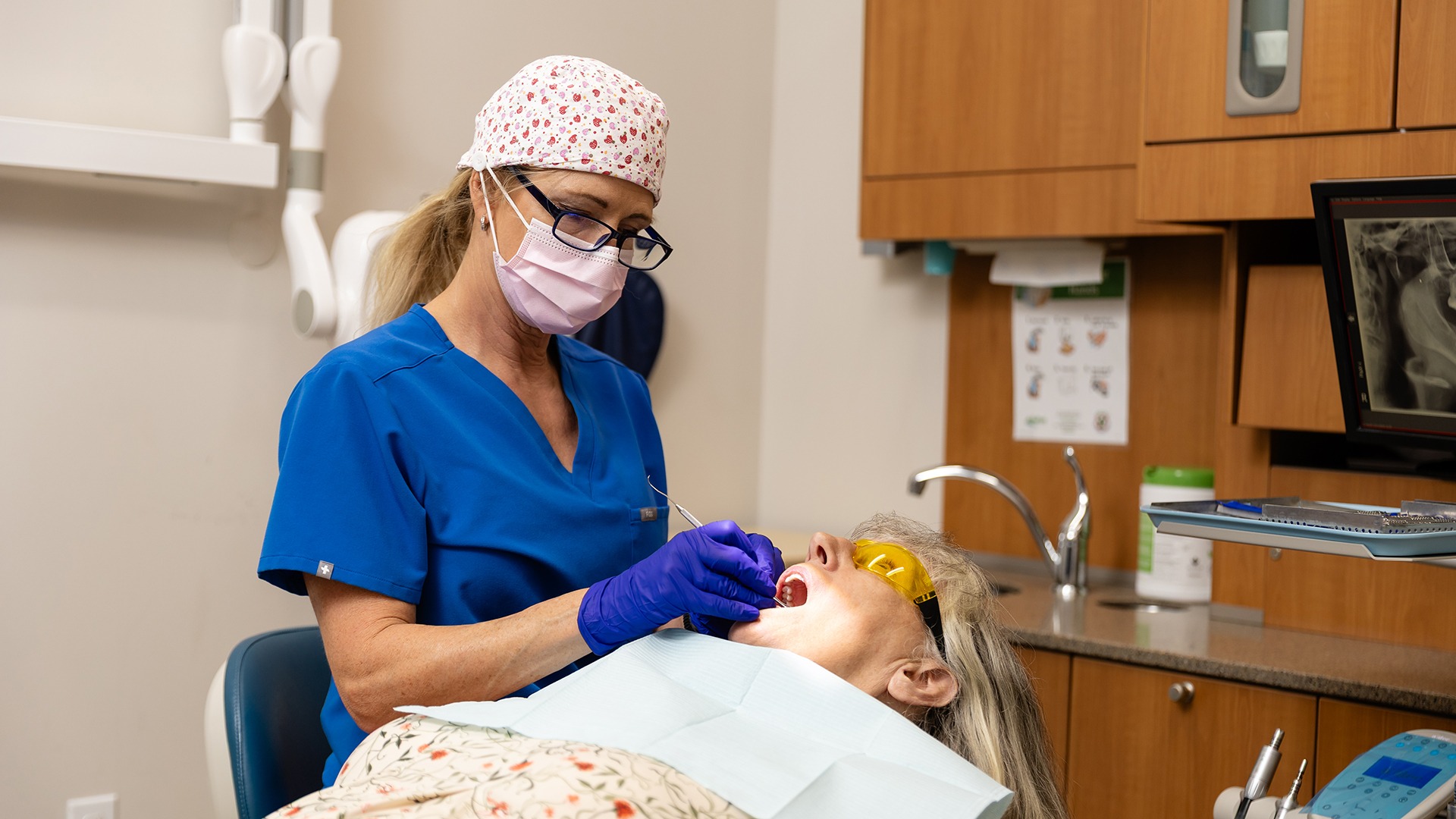 A person in scrubs, mask, gloves examines another person's teeth in a dental office with wood cabinetry and visible dental equipment.