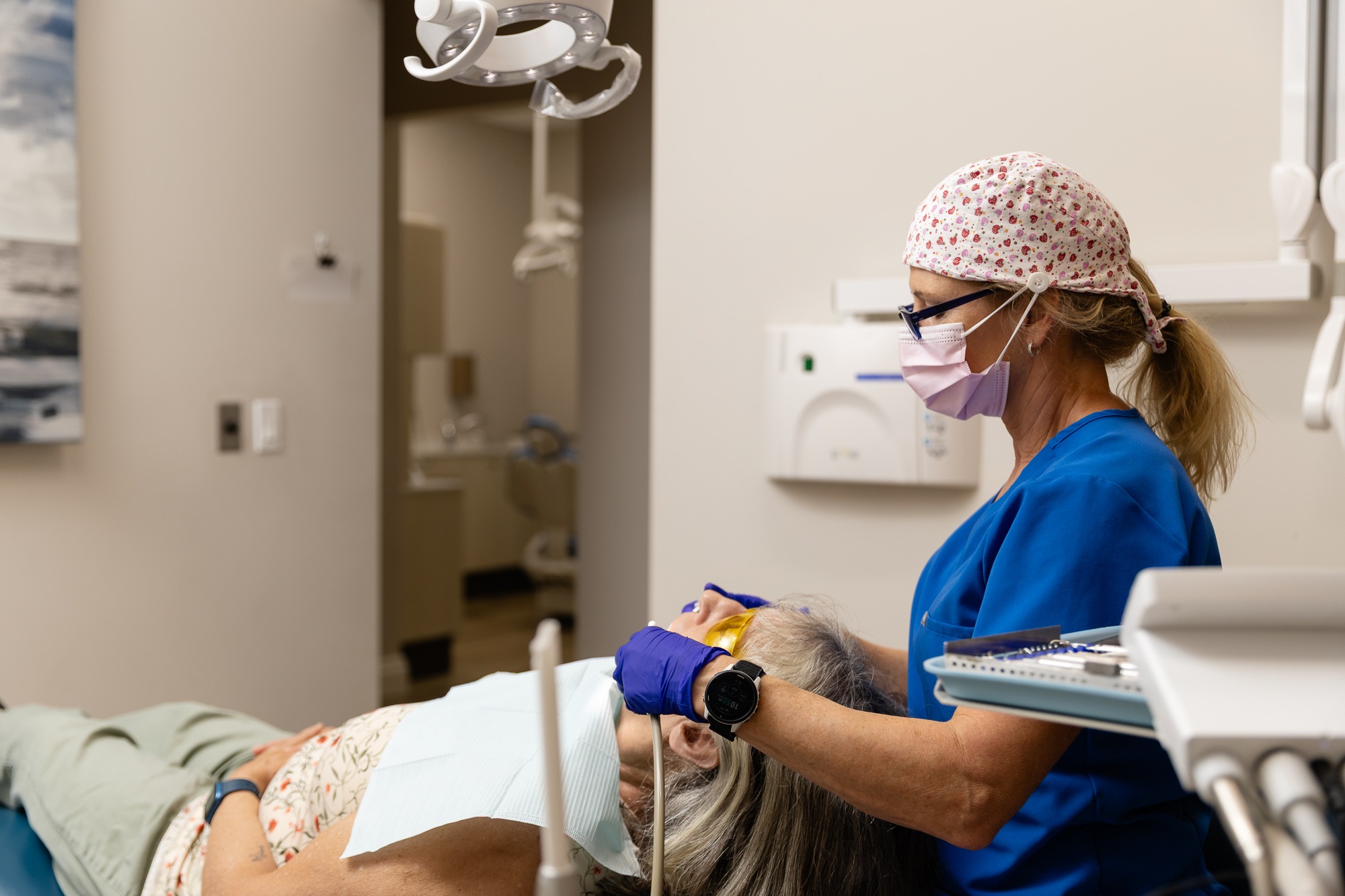 A person wearing scrubs and a mask examines a reclining person in a dental office, surrounded by medical equipment.