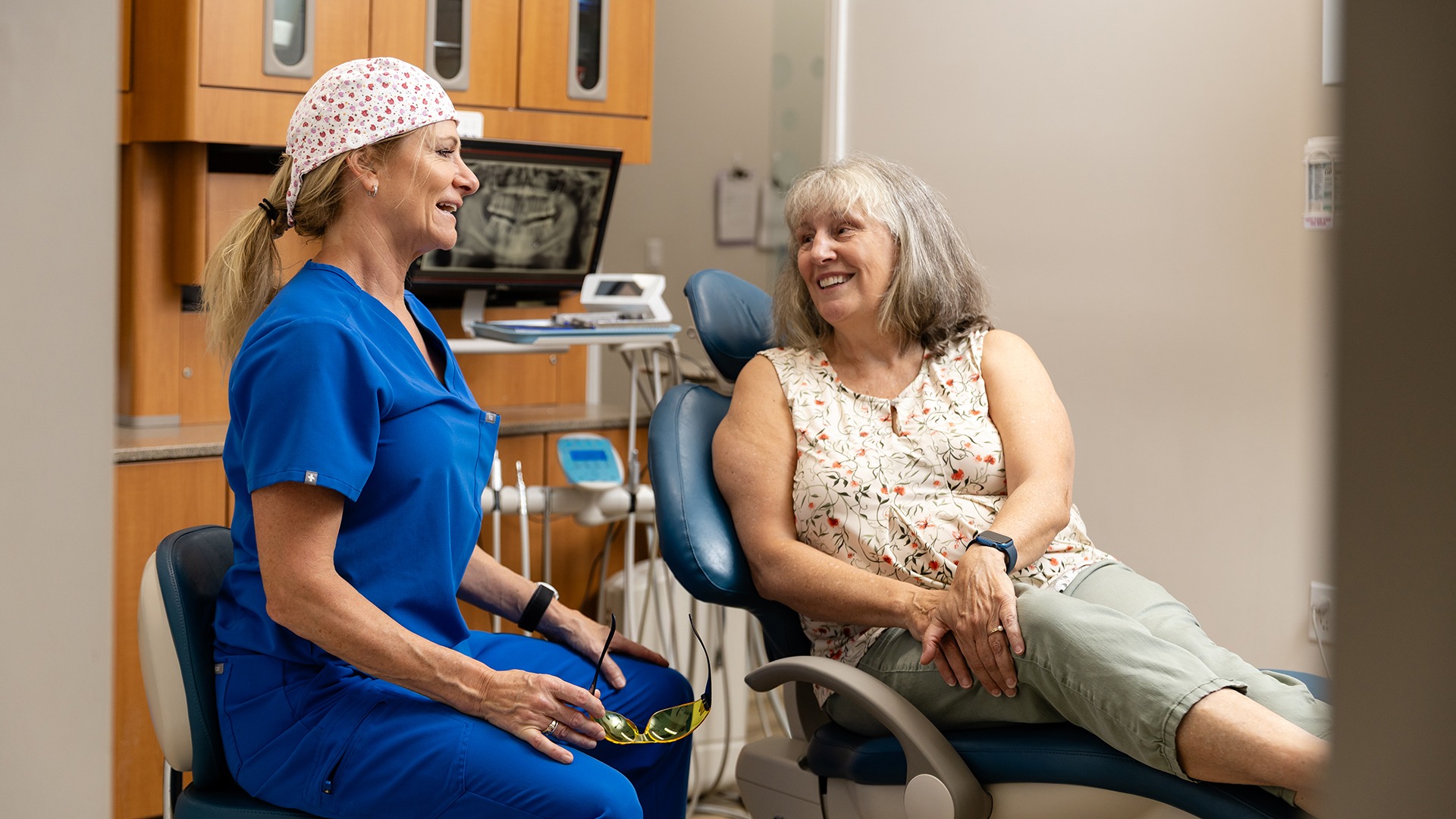 A person in blue scrubs converses with another person seated in a dental chair, inside a clinic setting.